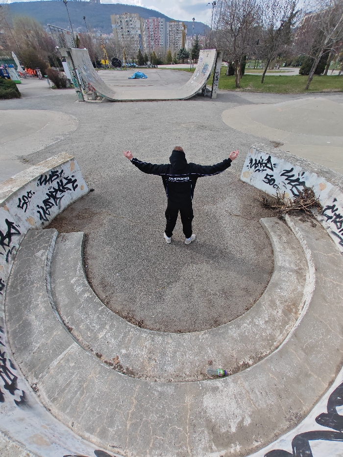 An aerial perspective image depicts an individual standing in a concrete skatepark within the city of Shumen, Bulgaria, specifically located in what appears to be a public recreation area or Youth Park, with the Shumen Plateau visible in the background.
The central subject is a person, viewed from behind, standing with arms horizontally outstretched and hands open. The individual wears a black hooded tracksuit with white horizontal stripes and "FACTORS" text across the upper back, paired with white athletic shoes. They are positioned on a grey, textured ground surface.
Immediately surrounding the person is a U-shaped concrete structure, characteristic of skatepark elements, featuring tiered ledges. This structure exhibits graffiti, including "50 ct." and other black spray paint markings on its inner walls. To the lower right on one of these ledges, a discarded clear plastic bottle with a green and purple label is visible.
In the midground, further behind the central figure, are larger concrete skate ramps or half-pipes, also showing signs of wear and graffiti. The ground throughout the skatepark area is a mix of worn concrete and a gravelly, dirt-like material. Bare, deciduous trees are sparsely distributed around the park perimeter, indicating a late winter or early spring season.
The background features several multi-story residential buildings, typical of post-socialist architecture, clustered in the middle distance. Beyond these structures, a large, tree-covered mountain or hill, identifiable as part of the Shumen Plateau, dominates the horizon under a partly cloudy sky. No other individuals are visible, and no direct interactions are depicted.