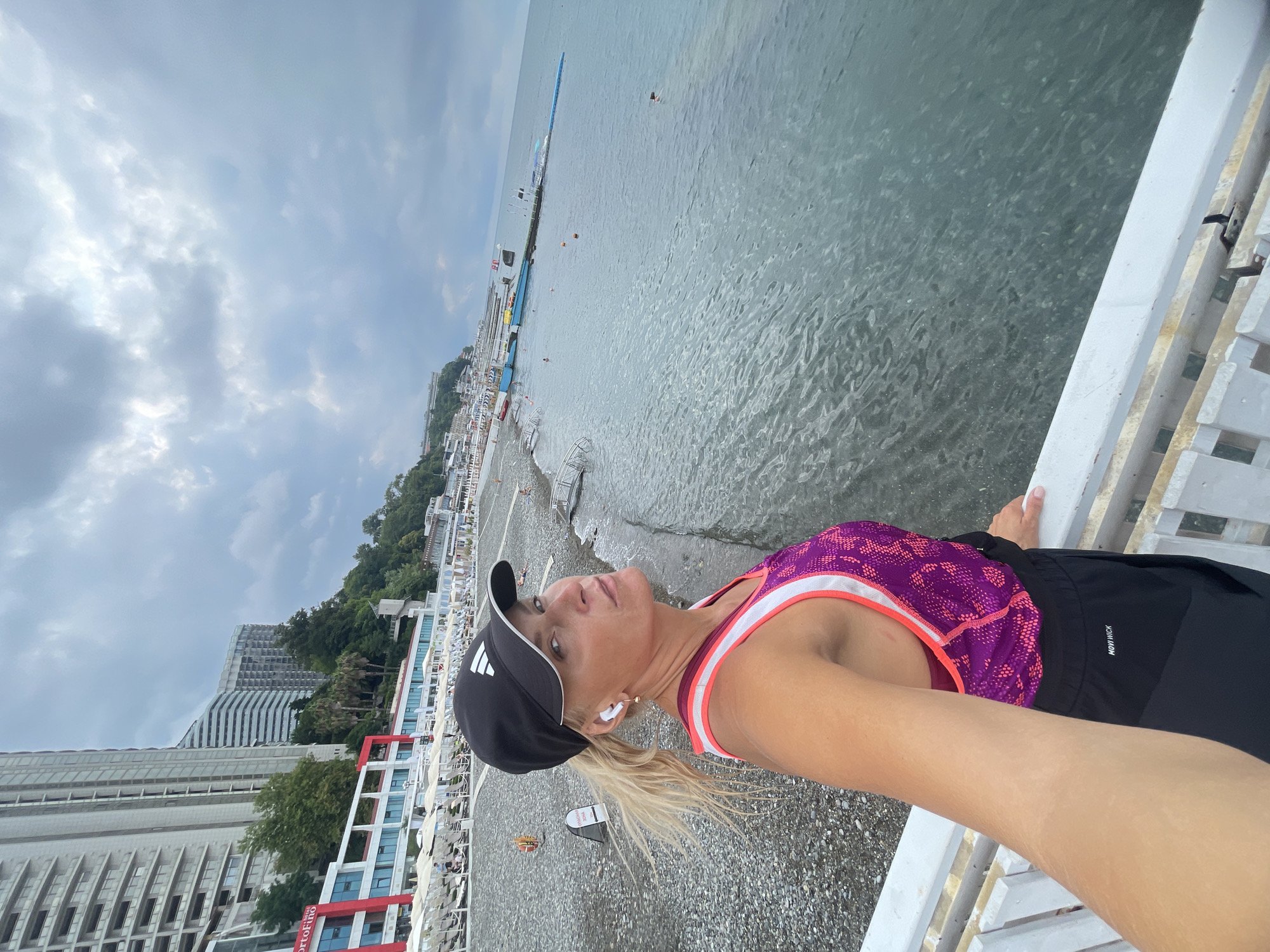 A blonde woman with fair skin is taking a selfie on a pier in Sochi, Russia. She is wearing a black baseball cap with white logos, white earbuds, a purple and pink athletic top with white trim, and black athletic shorts with white lettering. The pier has white railings. In the background, a pebbled beach is visible with lounge chairs and umbrellas. Beyond the beach are tall, modern buildings, some under construction, and lush green trees on a hillside. The sea is to the right of the woman, with a pier extending into the water, and buoys visible. The sky is overcast with dramatic clouds.