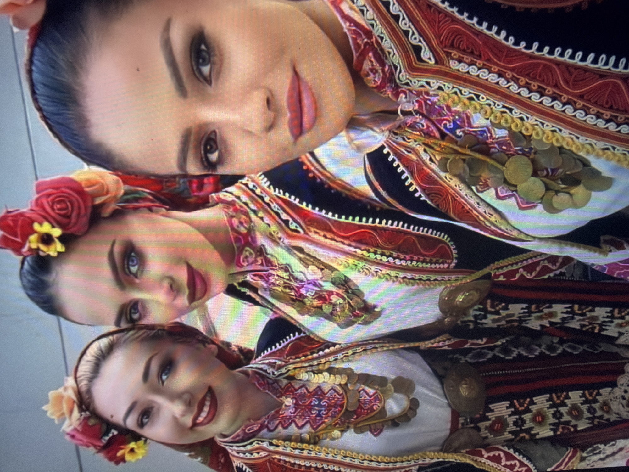 Three young adult women are depicted in a close-up, chest-up photograph captured in Shumen, Bulgaria. All three are attired in elaborate traditional Bulgarian folk costumes. They wear white long-sleeved blouses with intricate red and white embroidery around the neckline, topped with ornate vests or jackets featuring predominantly red, black, and white patterns with golden-yellow trim and detailed stitching. Each woman wears multiple strands of gold-colored coin necklaces cascading over their chests. The woman on the far left and the central woman also have large, ornate, gold-colored metal belt buckles visible at their waists. Their hair is styled and adorned with vibrant floral headpieces; the woman on the left has yellow and red flowers, the central woman features prominent red roses and yellow accents, and the woman on the right has red flowers. All three women wear visible makeup, including defined eyebrows, eye makeup, and red or pink lipstick, and are looking towards the camera with subtle smiles. The central woman exhibits distinct heterochromia or colored contact lenses, with her left eye appearing light blue/purple and her right eye appearing darker. The background is a plain, light-colored surface, exhibiting some horizontal light interference patterns.