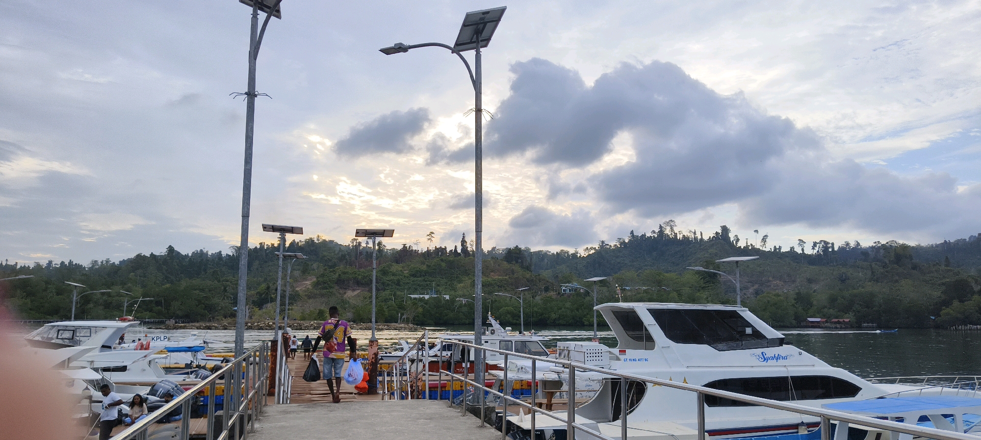 The image depicts a waterfront scene in Waisai, Indonesia, likely at a jetty or harbor. Several white speedboats are docked along a wooden pier, which is equipped with metal railings and streetlights. In the foreground, a man walks away from the camera, carrying two plastic bags. Another person is seated on the pier. The background features a lush green hillside covered in trees, with some buildings visible among the foliage. The sky is overcast with dramatic clouds, and the lighting suggests it is either early morning or late afternoon. The water in the harbor appears calm, with a small boat visible in the distance.