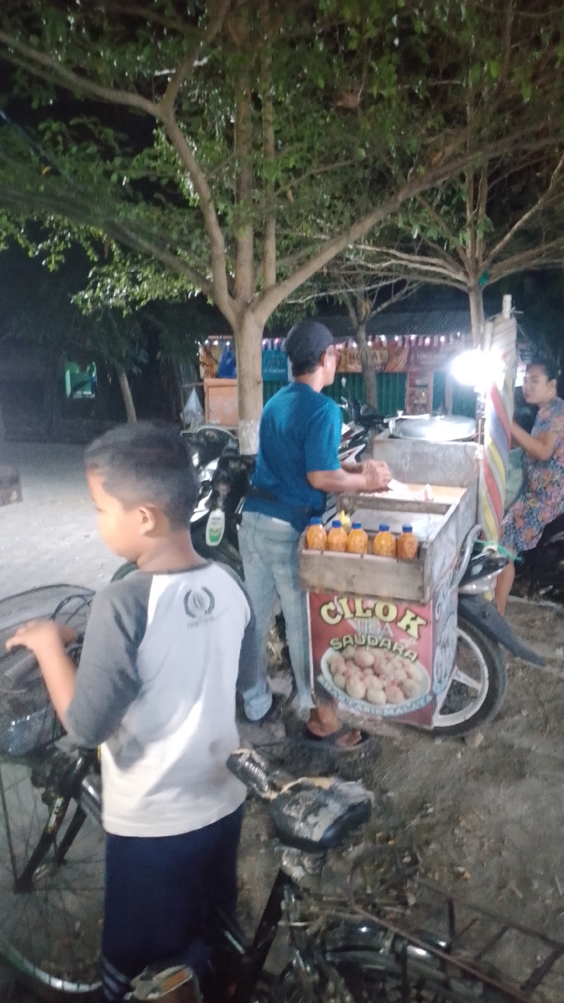 The image depicts a nighttime scene in the city of Karanglangu, Indonesia, at a street food stall. A young boy, seen from the side, stands in the foreground, wearing a grey and white t-shirt and dark blue pants. He is looking to his left. Behind him, a man in a blue t-shirt and light blue jeans is preparing food at a cart. The cart is decorated with a sign that reads "CILOK SAUDARA" and displays images of food items. Several bottles of sauce are lined up on the cart. To the right of the cart, a woman wearing a patterned dress is seated on a motorcycle. The environment appears to be outdoors with trees and some structures visible in the background, illuminated by artificial lighting. The ground is dirt or gravel.