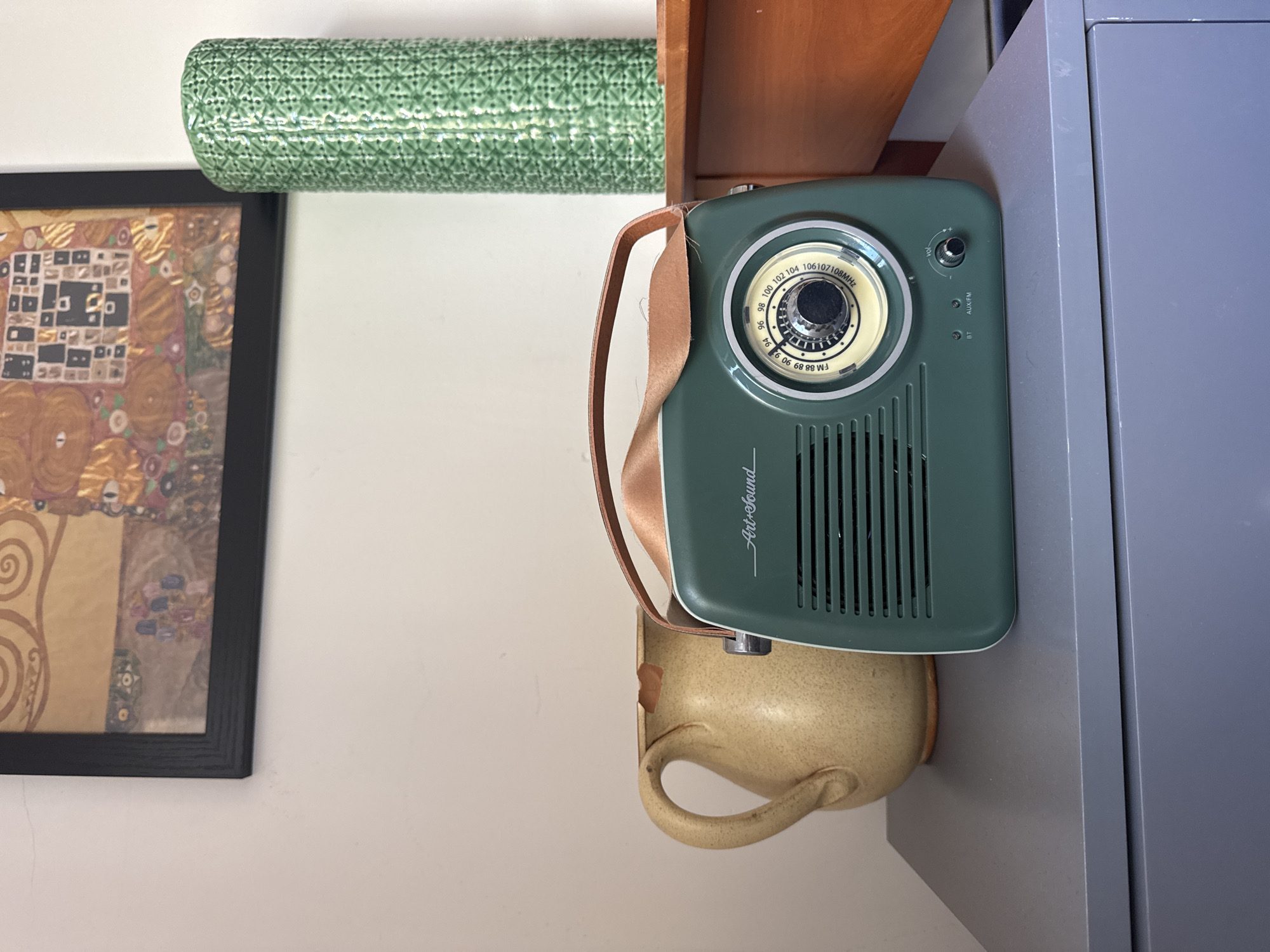 Here's a description of the image:
The image presents a collection of objects arranged indoors. On the left is a framed art print depicting a Klimt-style artwork with gold and geometric patterns. Above the print is a green cylinder, potentially a decorative item or ceramic piece.
To the right, resting on what appears to be a wooden shelf and a gray filing cabinet, is a vintage-style green radio with a tan carrying strap. The radio's dial is visible. Directly beneath the radio, a beige ceramic mug with a handle is partially visible.
The scene is likely indoors and the objects appear to be casually placed together. There is no evidence to suggest that this image was taken in London or the United Kingdom.