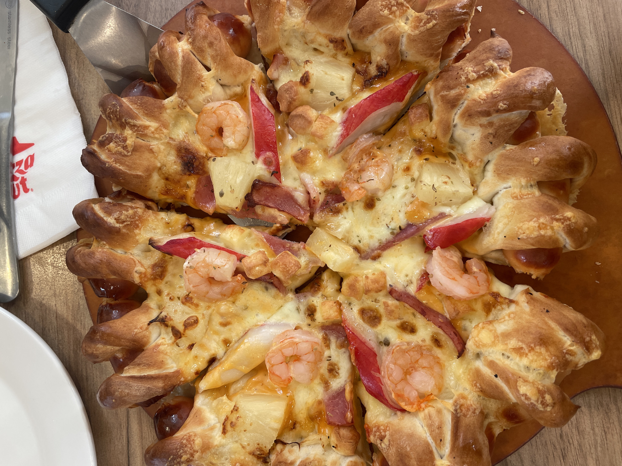 A close-up, overhead shot captures a freshly prepared pizza on a wooden table, likely in a restaurant setting. The pizza features a distinctive crust, intricately braided or formed into individual golden-brown rolls around the perimeter, some appearing to be stuffed. The top of the pizza is generously covered with melted, bubbly cheese, creating a rich golden surface.
Visible toppings include numerous plump, pink-orange cooked shrimp, bright yellow chunks of pineapple, and thin, reddish-pink strips which could be imitation crab or bell pepper. Smaller pieces of diced meat, possibly ham or bacon, are also scattered across the cheese. The pizza has been cut into several slices, with some slightly separated, suggesting it's ready to be served or has just started being consumed.
To the left of the pizza, a white paper napkin with the prominent red "Pizza Hut" logo is visible, indicating the restaurant chain. Resting on the napkin is a stainless steel pizza cutter or serving spatula, bearing the faint engraving "Stainless Steel". A portion of a white ceramic plate is visible in the bottom left corner, beside the pizza. The overall lighting is bright and even, highlighting the textures and colors of the food. The scene depicts a casual dining experience at a Pizza Hut, consistent with the provided location context of Tambon Fa Ham, Thailand.