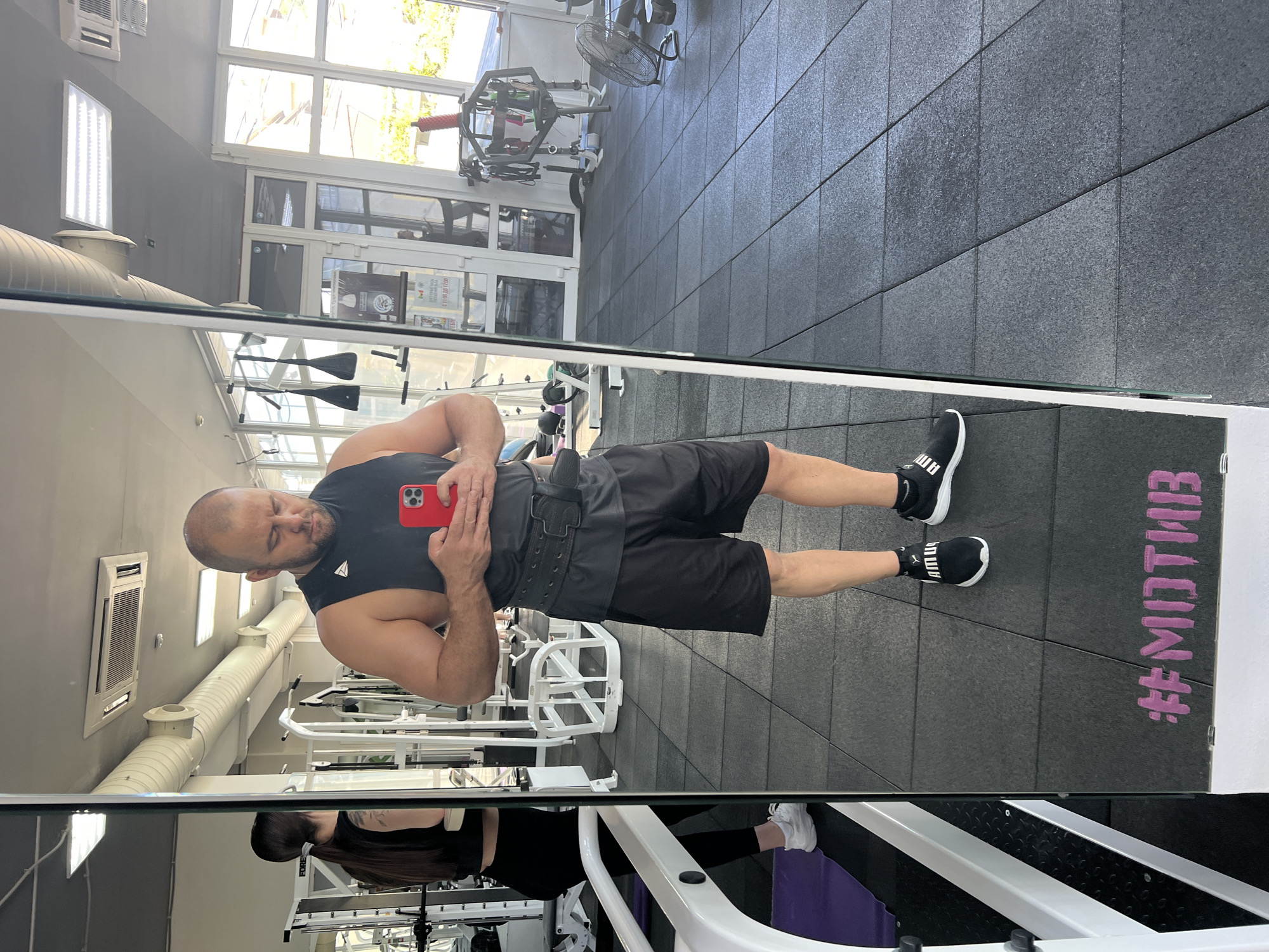 A bald man with a serious expression is captured in a mirror selfie inside a gym in Sochi, Russia. He is wearing a black tank top, black athletic shorts, and a black weightlifting belt around his waist. He holds a red smartphone with both hands to take the picture, his reflection prominent in the large mirror that dominates the left side of the frame. He wears black athletic shoes with white soles, and the "PUMA" logo is visible on his right shoe.
The gym features a dark gray tiled floor and extensive workout equipment, some of which is visible as white-framed machines in the background and foreground reflections. Bright natural light streams in from large windows along the back wall, suggesting it is daytime. Overhead, air conditioning units, fluorescent light panels, and a long white ventilation tube are visible.
Below the main subject's reflection in the mirror, another person, likely a woman, is partially visible, also dressed in black workout attire and white athletic shoes. Her reflection is also seen. On the dark gray floor mat, visible in the reflection, the stylized text "#МОТИВ" (Cyrillic for "#MOTIVE") is written in pink. The overall setting is a well-lit, modern fitness center.