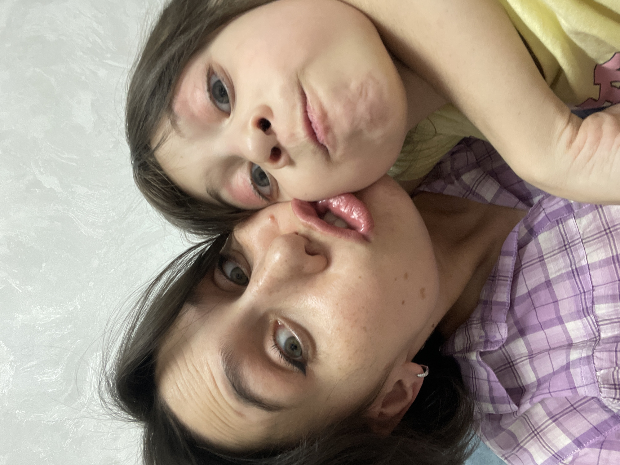 This close-up selfie features two individuals, an adult female and a young child, likely mother and daughter, captured in an intimate and playful moment indoors. The image is oriented diagonally, with both subjects looking towards the camera.
The adult female, positioned closer to the bottom of the frame, has dark hair, light skin, and light-colored eyes (green or hazel) accentuated with dark eyeliner. She sports a small silver hoop earring in her left ear and is wearing a purple and white plaid shirt. Her expression is animated and playful, with her mouth slightly open and her tongue visible, appearing to touch the child's chin.
The young child, positioned above and slightly behind the adult, has light brown hair, light blue-grey eyes, and fair skin. There are reddish marks or makeup around her eyes and on her cheeks. Her chin is resting against the adult's face, near her mouth. The child's expression is also playful, with her lips slightly pursed in a pout. She is wearing a light yellow top.
Their faces are in very close proximity, conveying affection and a lighthearted mood. The background consists of a plain, light-colored wall with a subtle texture, indicating an indoor setting. No specific time of day or weather conditions can be determined. There is no visible text in the image. The scene is located in Izhevsk, Russia.