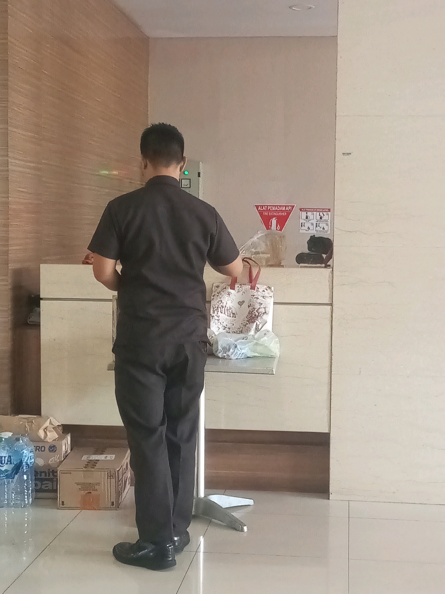 A man in a black uniform, likely a security guard, stands with his back to the camera in what appears to be an indoor public space. He is holding a patterned tote bag in his right hand and a clear plastic bag in his left. The man is wearing dark trousers and black shoes. In the background, there is a counter with a light-colored marble finish, and behind it, a wall with a wood-paneled texture. A fire extinguisher sign is visible on the wall to the right. To the left of the man, on the floor, are several boxes and bottles of water. The lighting suggests an indoor environment, possibly a lobby or reception area. The scene is static, with no apparent interaction between the man and any other individuals. The overall impression is one of a mundane, everyday occurrence within an urban setting, likely a commercial or institutional building in Bekasi, Indonesia.