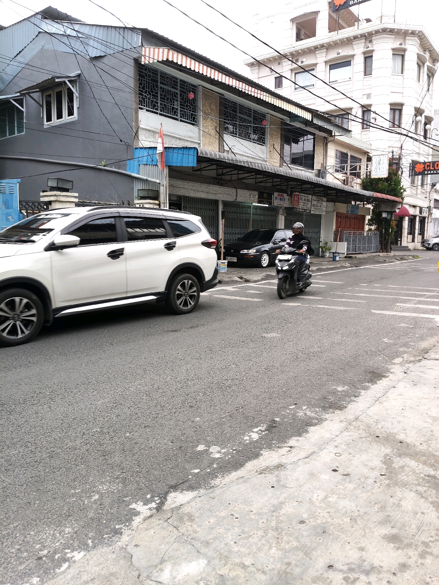In the city of Medan, Indonesia, on a paved road, a white SUV is parked on the left side of the frame, with its front wheels turned towards the right. A motorcyclist wearing a helmet and a dark jacket is riding a dark-colored scooter across the intersection, moving from left to right. The road has white lane markings. In the background, a multi-story building with a light-colored facade and multiple windows stands on the right. To the left of this building, a narrower, darker-colored building features a red and white striped awning and several windows. An Indonesian flag is visible on a pole in front of the darker building. Several electrical wires are strung across the scene, obscuring parts of the buildings and sky. The environment appears to be a commercial or mixed-use urban area.
