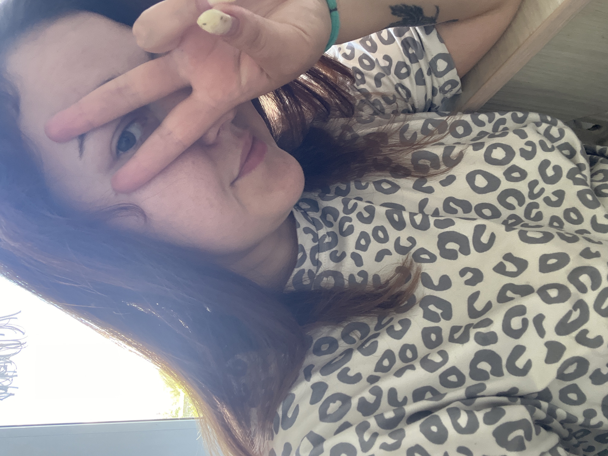 The image is a selfie of a young woman with long, brown hair. She is lying down, with her head tilted and her right hand raised to her face, making a peace sign. Her left arm is resting on a wooden surface. She is wearing a white shirt with a grey leopard print pattern. Her left eye is visible, and she has a slight smile. There is a tattoo visible on her upper left arm, and a turquoise bracelet on her wrist. The lighting suggests it is daytime, with natural light coming from the left side of the frame. The background is blurred, but appears to be indoors with some hints of outdoor scenery visible through a window or opening. The overall mood appears casual and relaxed.