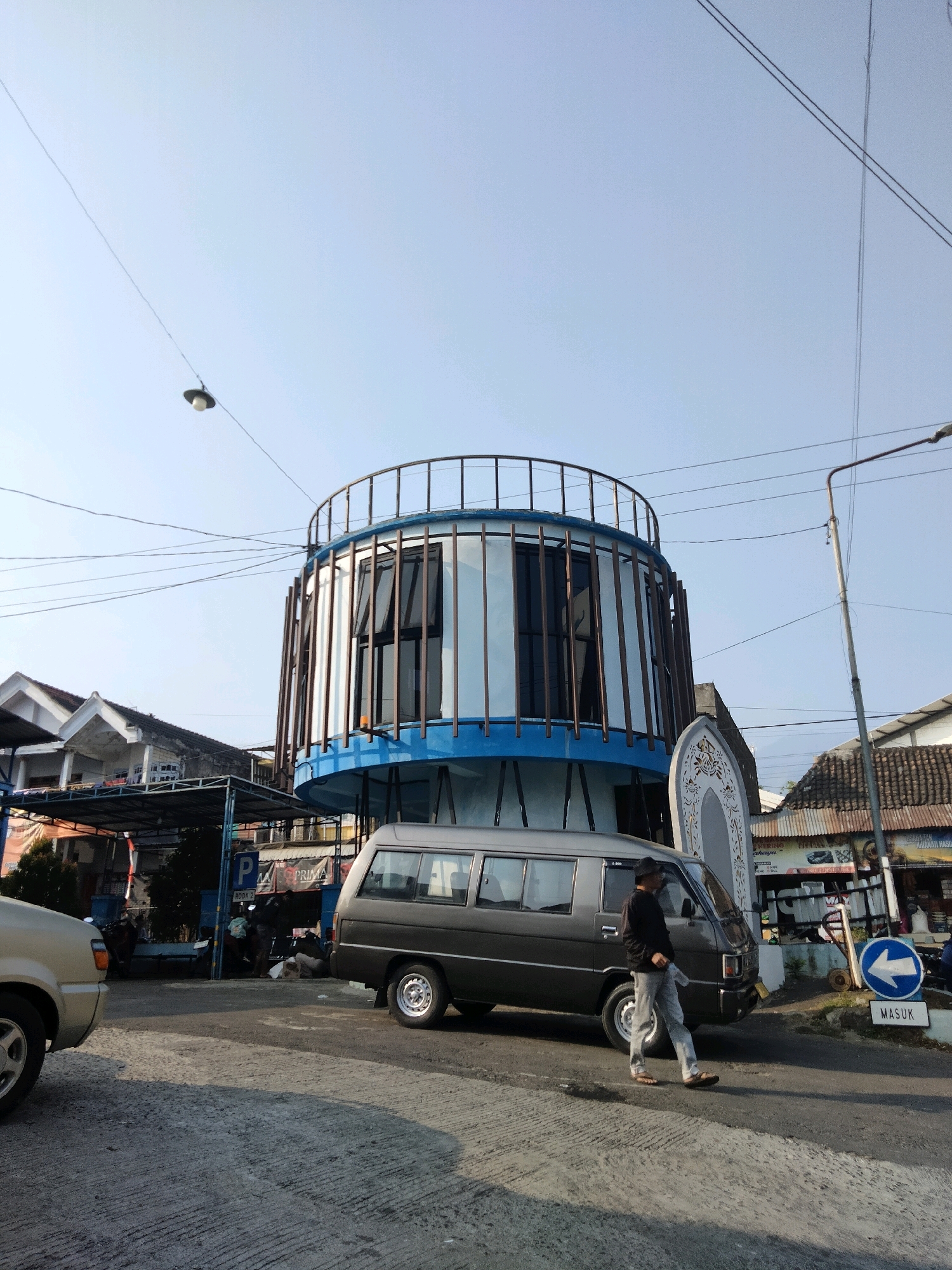 In Sawahan, Indonesia, a gray minivan is parked on an asphalt road, with a man walking in front of it. The man is wearing a black long-sleeved shirt, light-colored pants, and sandals. He is holding a clear plastic bottle. The minivan has its headlights on and the parking brake engaged. In the background, a distinctive circular building with a blue and white facade and vertical brown supports is visible. To the left of the minivan, a portion of a gold-colored car is parked. Further in the background, there are buildings with tiled roofs and a street lamp. A blue directional sign indicating a left turn and a sign reading "MASUK" (enter) are also present. The sky is clear and blue.