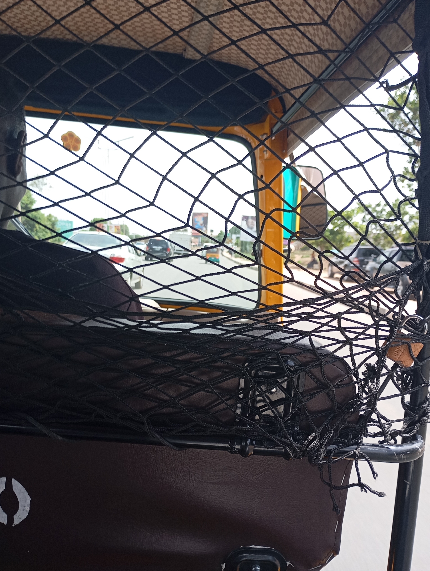 The image provides a view from inside a three-wheeled auto-rickshaw, commonly known as a Keke Napep, looking forward through a prominent black mesh safety net. This net forms a grid pattern across the entire frame. In the immediate foreground, beneath the net, a brown upholstered panel, possibly part of a seat or storage compartment, displays a white painted symbol resembling a stylized eye or an inverted 'U'.
On the left side of the frame, the back of a person's head is visible, indicating they are positioned in front, likely driving or occupying the front seat. The interior of the vehicle features yellow and turquoise panels and a beige, patterned roof lining. A small, yellowish, paw-shaped ornament is attached to the mesh net near the top left, and a keychain with keys hangs from the net structure on the right.
Beyond the vehicle's interior, a daylight street scene unfolds. Several cars, including a white sedan and darker SUVs, are visible on what appears to be a paved road, suggesting the vehicle is in traffic or on a busy route. Trees and green vegetation line the sides of the road in the mid-distance. Faint, unreadable text can be seen on billboards further in the background. The bright lighting suggests it is daytime. The scene depicts a typical urban or semi-urban setting in Jos, Nigeria.
