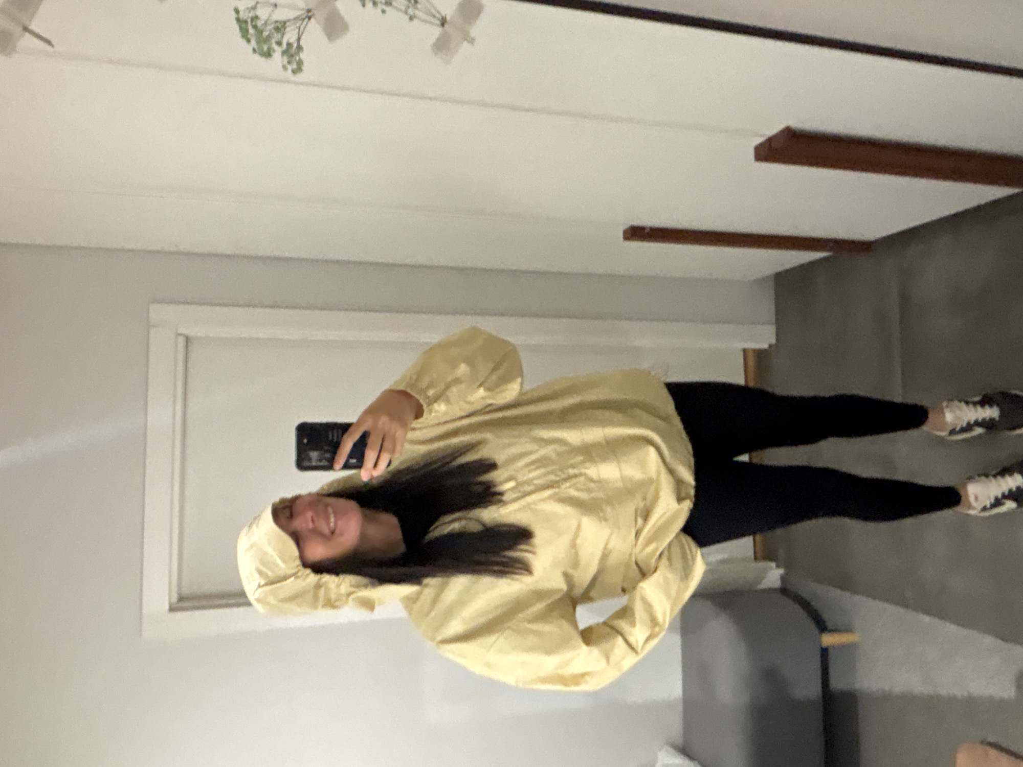A person is indoors, taking a selfie in what appears to be a room. The person is wearing black leggings and a light yellow hooded jacket. They are holding a smartphone up to take the picture. The room has white walls and ceiling, with a doorframe visible. There is a light fixture on the ceiling. A grey stool is positioned on the floor. Based on the location in the city of Chaikovsky, Russia, the environment is likely a residential setting.