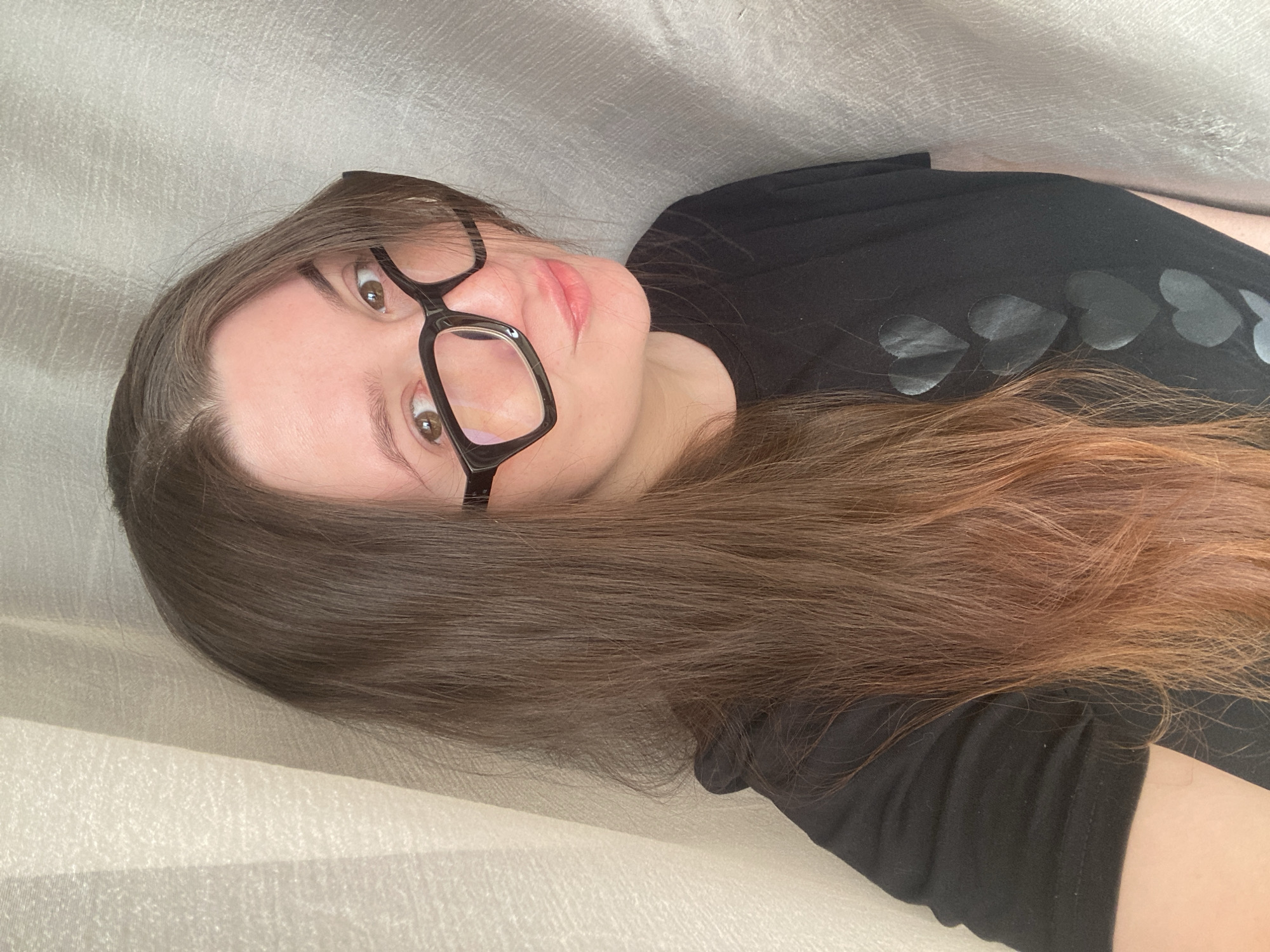 A woman with long, brown, slightly ombré hair is lying down on what appears to be a textured, light-colored surface, possibly fabric or a bed covering. She is wearing black-framed glasses and a black t-shirt with a pattern of matte black hearts on the sleeve. Her gaze is directed toward the viewer.
There is no information in the image to support the location in the city of Chaikovsky, Russia.