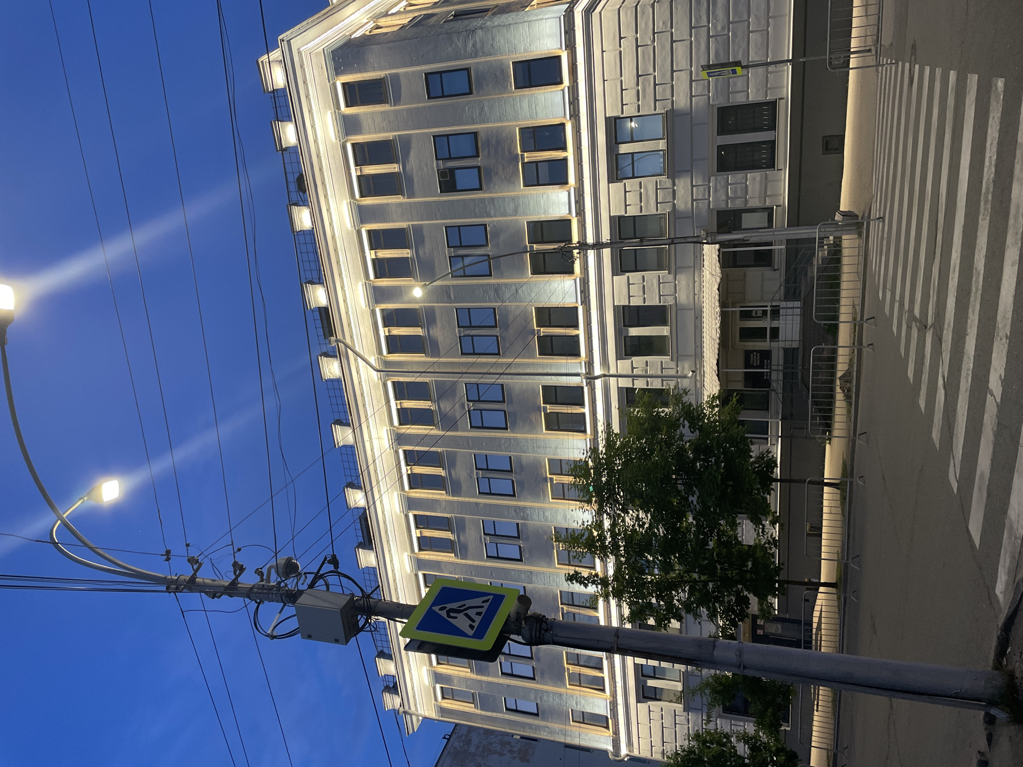 The image captures a nighttime street scene in Vologda, Russia, likely featuring a pedestrian crossing on a city street in front of a multi-story building. The building, illuminated by external lights, exhibits a classical architectural style with decorative elements along its facade and rooftop. A street lamp extends from a pole in the foreground, casting light on the scene. The pole also supports a pedestrian crossing sign and what appears to be a surveillance camera and electrical equipment. Several trees line the sidewalk in front of the building, and metal barriers are present along the street. The sky is dark blue, indicating it is either dusk or night. The overall impression is one of urban infrastructure and public safety measures within a city setting.