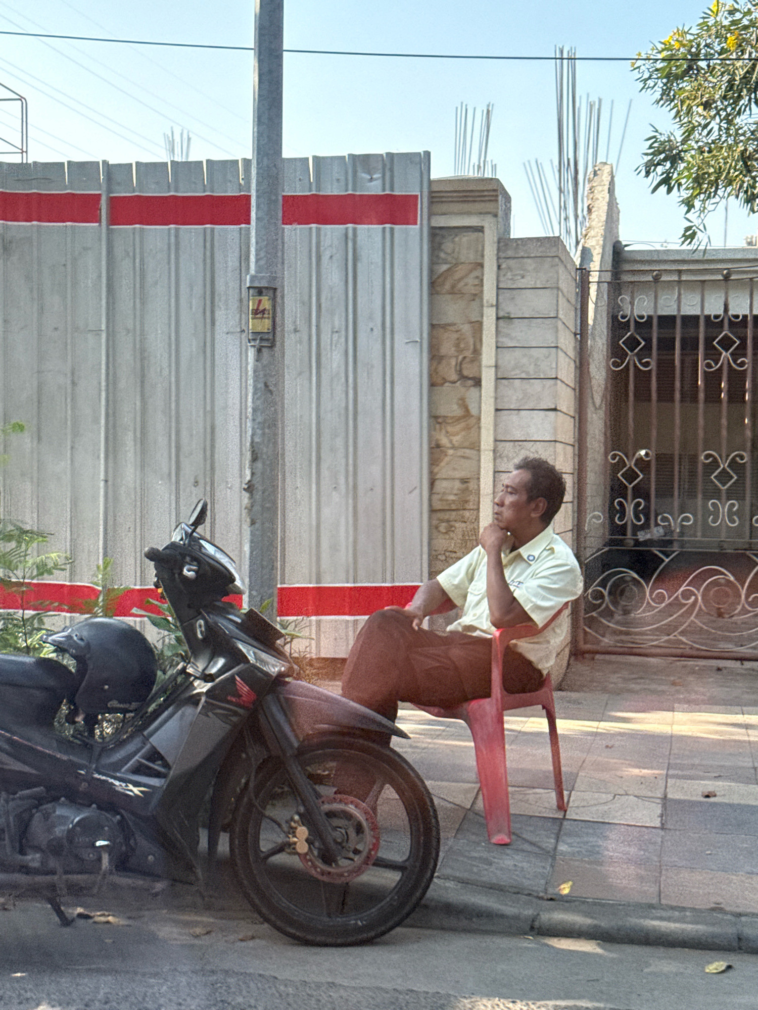 A man wearing a light yellow short-sleeved shirt and dark brown pants sits on a red plastic chair on a sidewalk. The man is looking to his left, with his chin resting on his hand. He appears to be in a contemplative or relaxed posture. To his left, a black motorcycle is parked. The motorcycle has a black helmet on its handlebars. Behind the man and the motorcycle is a textured concrete wall with a red horizontal stripe. Further in the background, there are ornate metal gates and the top of a building with visible construction rebar. The scene suggests an urban environment, likely in Surabaya, Indonesia, given the style of architecture and the presence of a motorcycle, a common mode of transportation in the region. The lighting indicates it is daytime, with sunlight casting shadows on the ground.