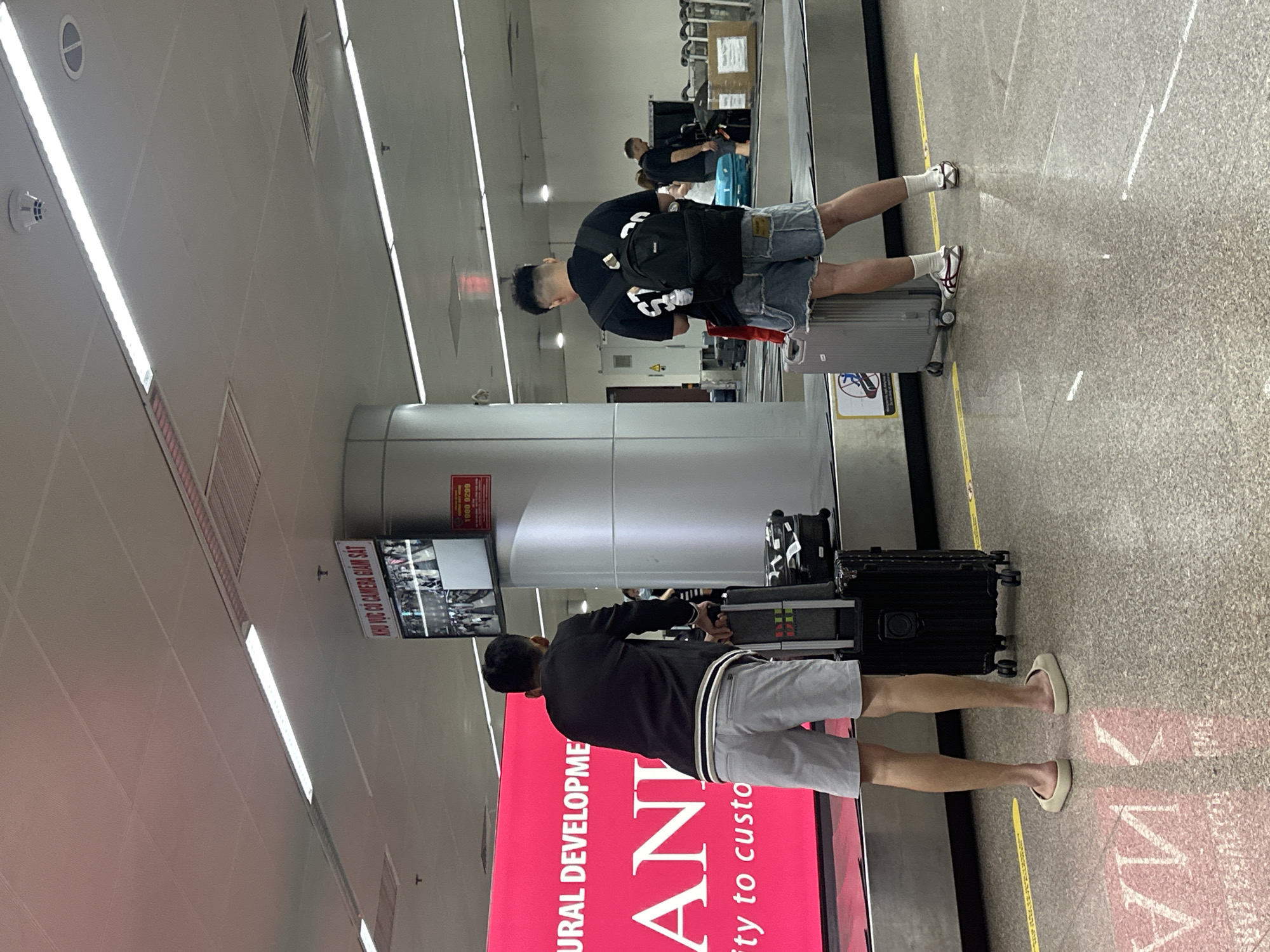 The image shows the baggage claim area of an airport, potentially in Đà Nẵng, Vietnam, based on the text on a nearby advertisement. Two men are present, each with luggage. One man wears a black shirt, denim shorts, and sneakers and is retrieving a gray suitcase from the baggage carousel. The other man, closer to the camera, wears a black jacket, gray shorts, and flip-flops; he also has a suitcase. Both individuals appear to be waiting for or handling their luggage. The area has overhead lighting, safety yellow lines on the floor, and a display screen stating "KHU VỰC CÓ CAMERA GIÁM SÁT" (Area Under Camera Surveillance) and a phone number. Adjacent to the man closer to the camera is a pink advertisement with the words "URAL DEVELOPMENT" and "ANI."