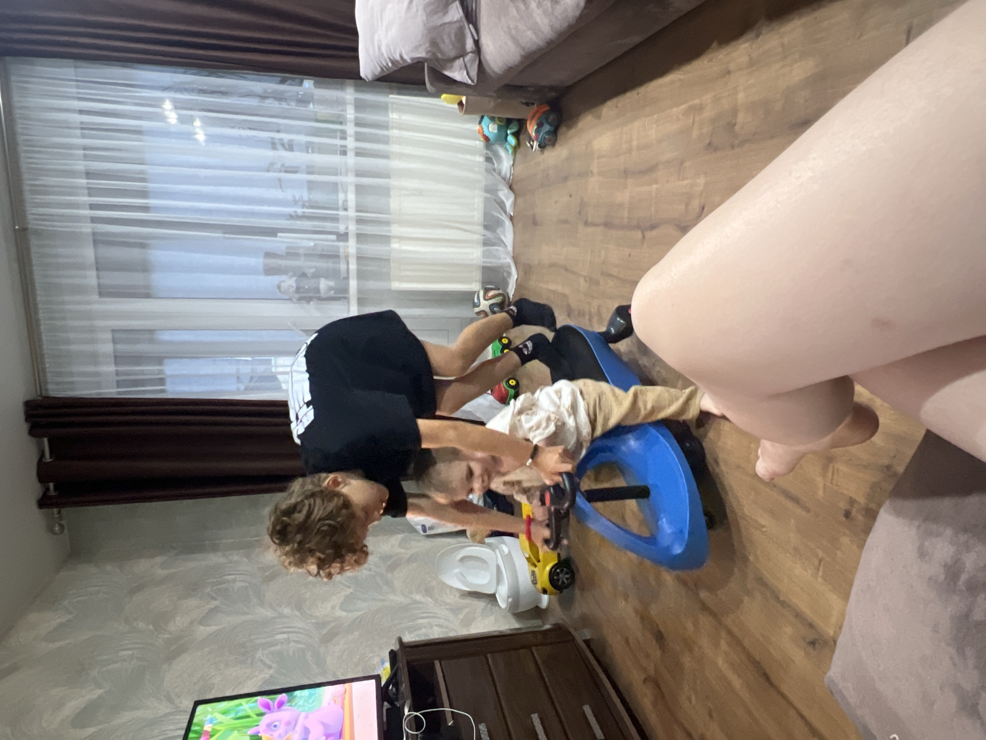 In a living room in Noviy, Russia, a young child, dressed in a light-colored shirt and pants, sits on a blue toy car. An older child, wearing a black t-shirt and dark socks, leans over the younger child, appearing to assist or play with them. The children are on a wooden floor. To the left, a dresser with drawers is visible, with a television screen showing a cartoon character. Behind the children, a white and gray patterned wallpaper adorns the wall. A brown curtain hangs on the right side of the frame, and sheer white curtains cover a window behind them. In the background, a sofa is partially visible, and various toys are scattered around the room, including a yellow toy car, a small red toy, and a blue octopus toy. The scene appears to be a home environment.