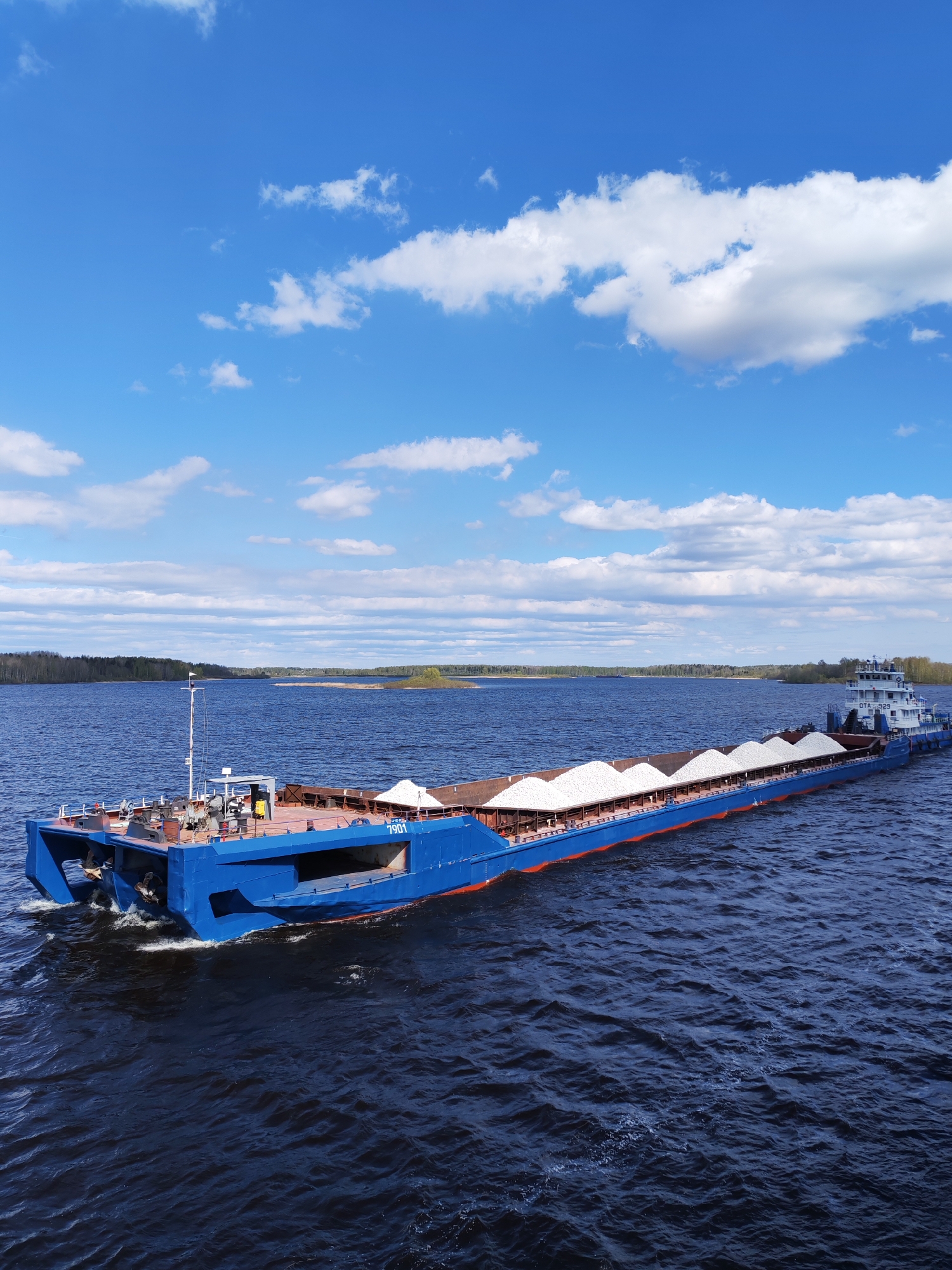 A wide-angle shot depicts a blue barge heavily laden with a cargo of white material, navigating a large body of water. The barge is primarily blue with a red accent along the middle. The cargo appears to be piled into several cone-shaped mounds. The water is a dark blue with slight ripples. In the background, there is a strip of land covered with trees. The sky is a vibrant blue with scattered white clouds. The location may be near Pochinok, Russia, given the presence of similar barges in the area. The primary subject is the barge in motion, transporting its cargo across the water.