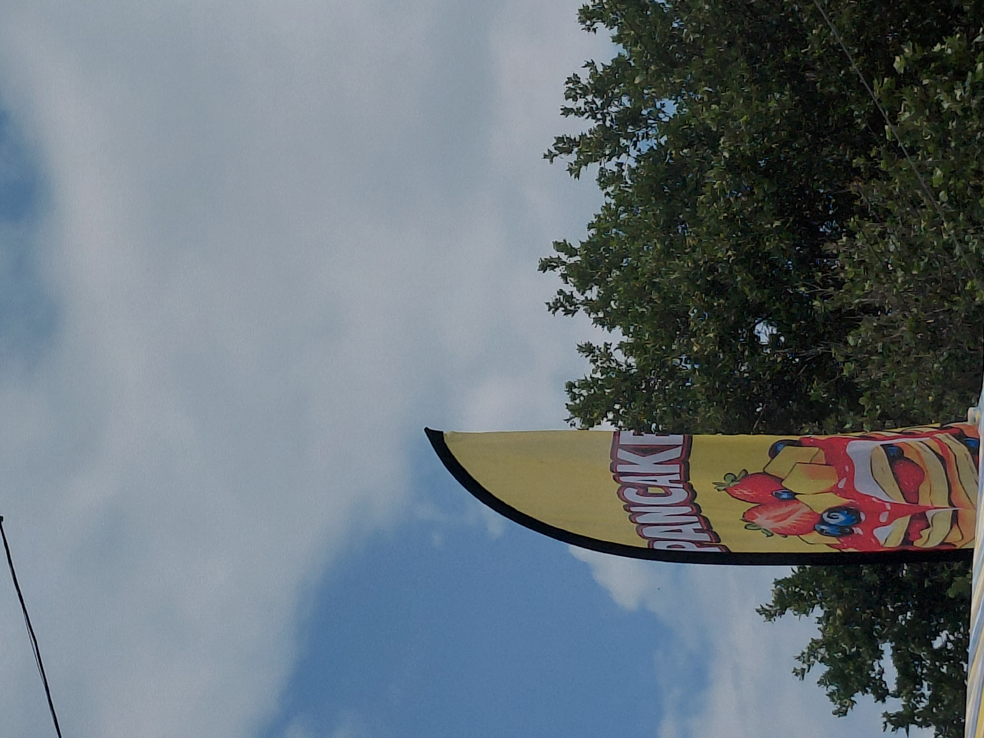 A vertical, yellow feather flag, outlined in black, dominates the central-left portion of the image. The flag displays the partially visible word "PANCACKE" in red letters with white outlines, situated above an illustration of a stack of pancakes. The pancakes are depicted with red syrup cascading over them, topped with red strawberries, blue blueberries, and yellow diced fruit. The flag is positioned outdoors against a backdrop of a partly cloudy blue sky, featuring white and light gray cumulus clouds. Dense, dark green foliage from deciduous trees occupies the right side and lower background of the frame. A thin, black wire or cable stretches diagonally across the upper left corner of the image. No individuals are visible in the scene. The setting is in the city of Kostievo, Bulgaria.