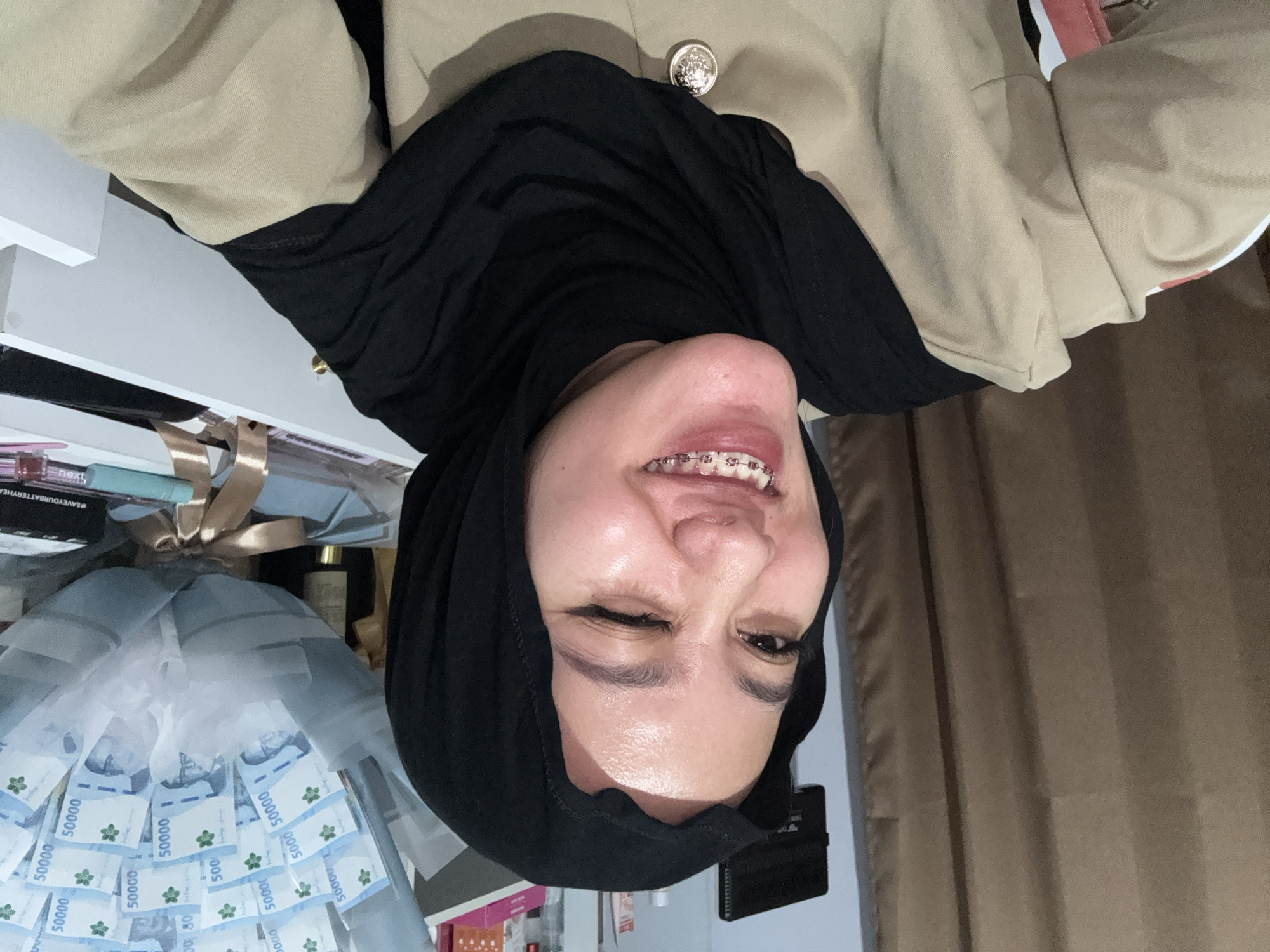 A young woman wearing a black hijab and a tan jacket is pictured in a selfie, winking and smiling. She has braces on her teeth. Behind her, a bouquet of money is visible, alongside various items on shelves and in drawers, including what appears to be a black electronic device on the wall. The background also includes a brown curtain. The setting is indoors, likely a room within a residential or commercial space in Gadingrejo, Indonesia.