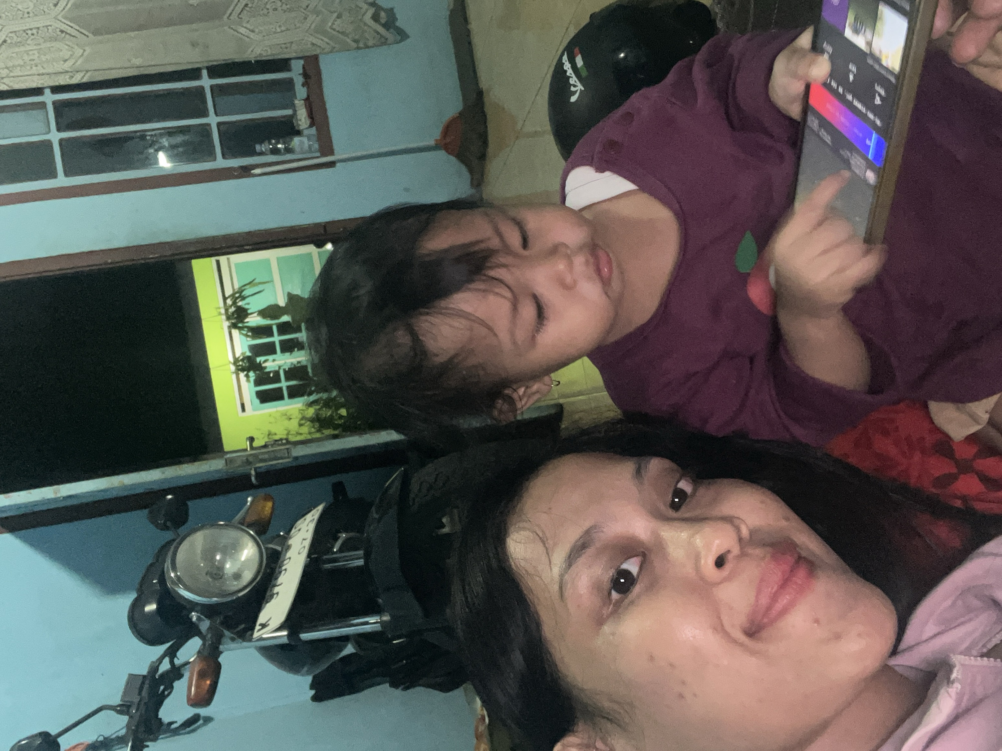 The image presents a close-up, upward-angled view of a woman and a young child in what appears to be an indoor or covered outdoor setting during the evening or night. The overall impression is one of a casual, shared moment.
In the foreground, a woman with long, dark hair and a fair complexion is seen from the chest up, looking directly at the camera with a relaxed expression and a slight smile. She is wearing a light-colored, possibly pink or light purple, top.
Above and slightly behind her is a young child, likely a toddler or preschooler, dressed in a dark purple long-sleeved top with a small red and green emblem on the front. The child is holding a smartphone with both hands, intently looking at and interacting with the screen, which displays a colorful user interface with red, blue, and purple gradients and various icons. A black helmet with a white "Vespa" logo is visible near the child's head.
The background features architectural elements: a light blue painted wall spans the left side and top, with an intricately patterned ceiling border. Through a window or opening in the blue wall, a brightly lit, green-painted building or wall with its own window panes and some green foliage is visible. Below the visible wall, parts of a motorcycle are seen, including a chrome-ringed headlight, an amber turn signal, and a white license plate with the partial text "KT 54** Y" and the date "07-25" (July 2025). The dark surroundings suggest it is nighttime, with artificial light illuminating the scene.
The image captures a familial interaction, possibly a mother and child, engrossed in a shared activity involving a smartphone, set in a domestic environment in Kecamatan Pati, Indonesia.