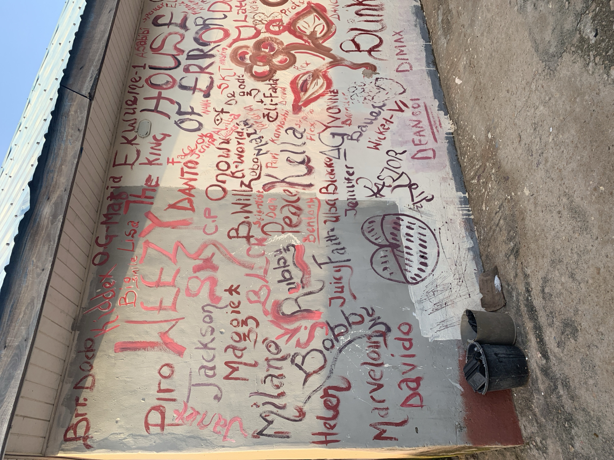 The image depicts a section of a building's exterior wall in Jalingo, Nigeria, covered in graffiti. The wall is painted a light gray and is marked with various names and phrases in red and dark purple paint. Some of the visible text includes "Brr. Dodo," "Piro," "WEEZY," "Jackson," "Maggie," "Milano," "Helen," "Marvelous," "Davido," "The KING HOUSE OF ERROR," "B. Willz," "Scientist," "Peace," "Kella," "Juicy Faith," "Uba Blacko," "AGYVONNE," "Barrister," "Wicked-VI," "KASTOR," "DIMAX," and "DEAN COI." There are also drawings, including a stylized heart shape and a flower. The base of the wall is painted red, and at the bottom left, two dark-colored trash bins are visible on the ground, one filled with black objects and the other with black bags. The ground is rough and appears to be a dirt or gravel surface, with some white paint splatters visible. The building has a metallic roof. The sky is visible in the upper right corner, appearing clear and blue. The graffiti suggests a public space where individuals have inscribed their names and potentially messages.