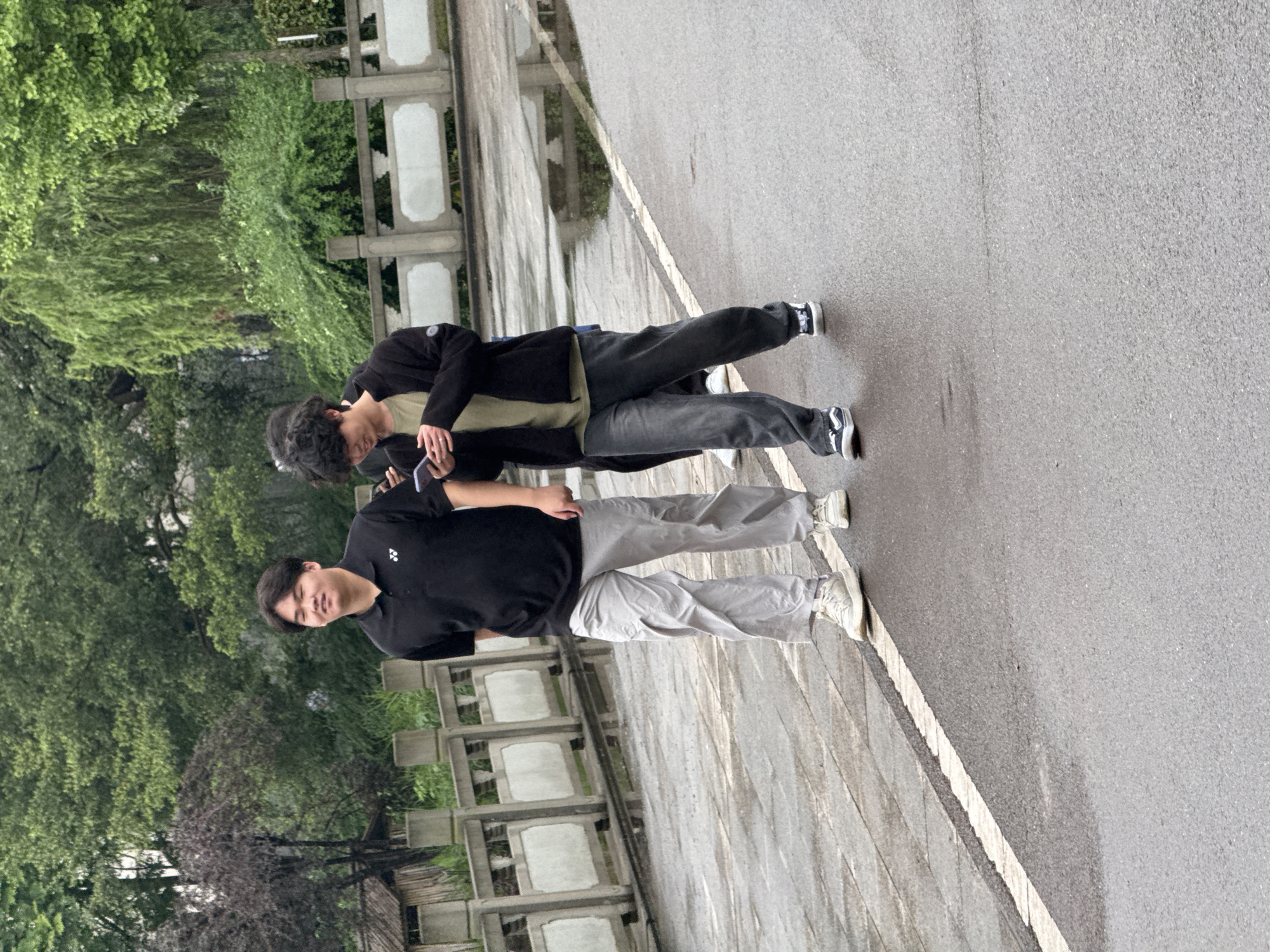 Two people stand side by side on an asphalt path, with a white line marking the edge of the pavement. The person on the left wears a black polo shirt and light gray pants, while the person on the right wears a black jacket over a light green shirt and dark pants. Trees and greenery line the left side of the path, and a row of decorative concrete structures borders the paved area. The location is likely Jiangjin, China, based on visual elements such as architecture and vegetation.