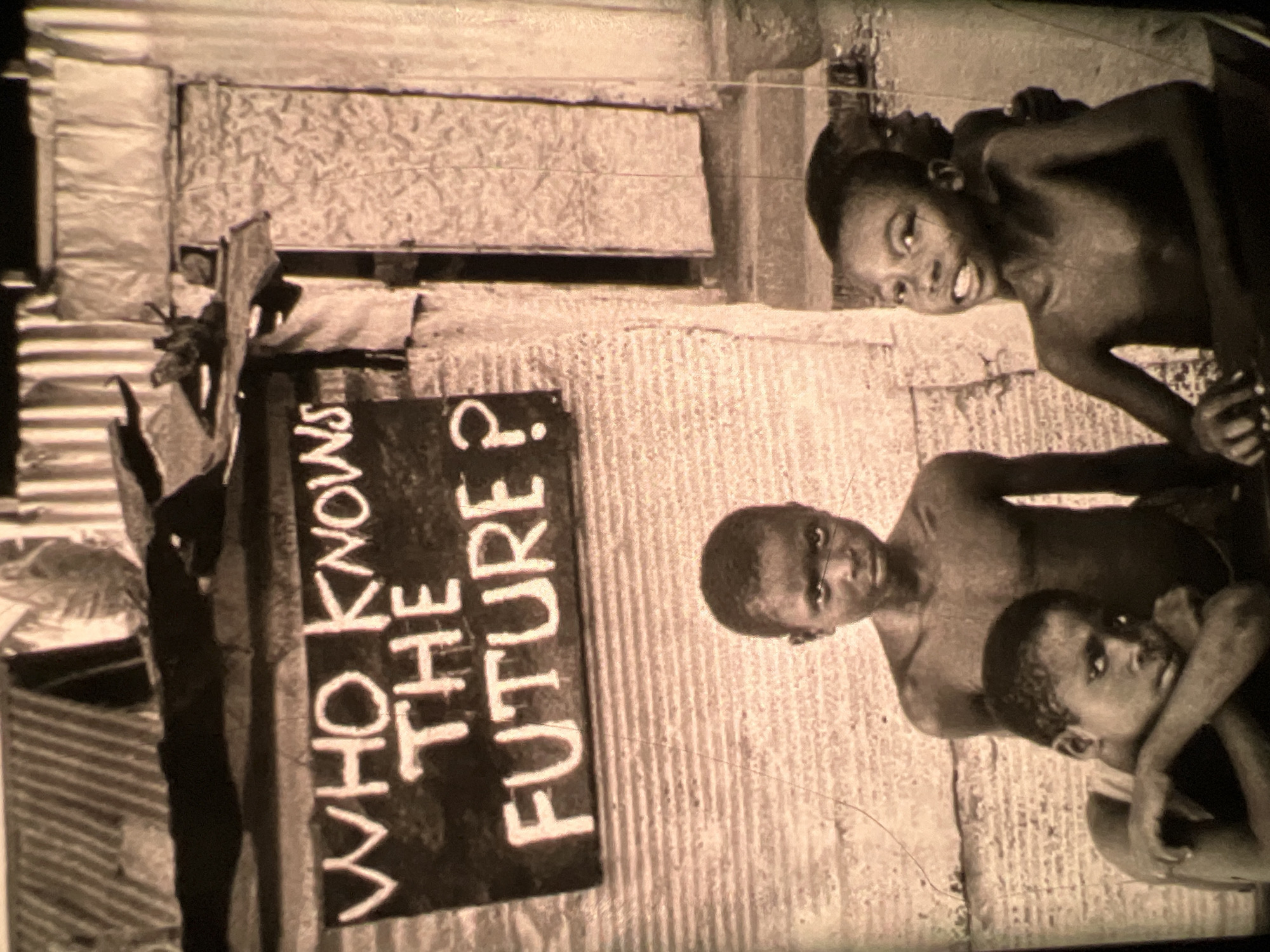 In the city of Lagos, Nigeria, a sign with white lettering reads "WHO KNOWS THE FUTURE ?" against a dark background. Below the sign, three young boys are gathered. The boy on the left, in the foreground, rests his head on his arms, looking towards the viewer. Behind him, a second boy stands, also looking at the viewer with a somber expression. To the right, a third boy smiles and looks forward, his body slightly angled. The environment appears to be an outdoor setting, possibly a street or alley, with makeshift structures and corrugated metal visible in the background. The overall impression is one of poverty and uncertainty, conveyed through the expressions of the children and the questioning sign.