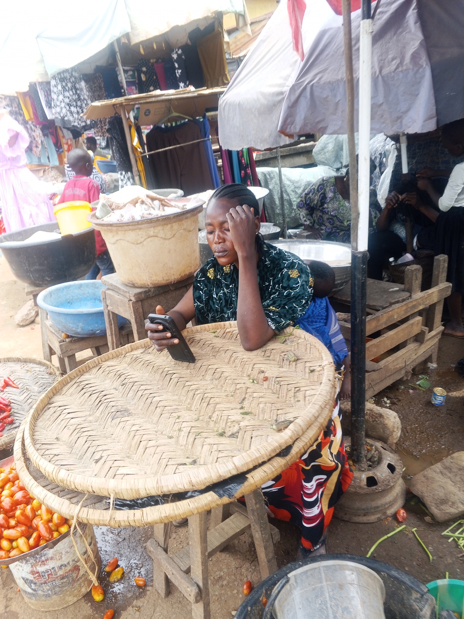 In a bustling outdoor market in Kabba, Nigeria, a woman is seated at a rustic wooden table, her gaze fixed on her mobile phone. She wears a dark patterned blouse and a vibrantly colored wrapper. A baby is secured to her back with a blue cloth. In front of her, a large, woven tray rests on the table, laden with dried leaves. To the left of the table, a metal bucket overflows with ripe red tomatoes. Further left, a dark basin holds a white substance, possibly rice or flour, and a smaller blue basin sits below it. In the background, market stalls display a variety of clothing and fabrics, with awnings providing shade from the sun. Several people can be seen browsing or working in the market. The ground appears to be dirt and mud, indicative of a humid climate. The overall scene depicts a typical day at a Nigerian market, characterized by commerce, community, and daily life.