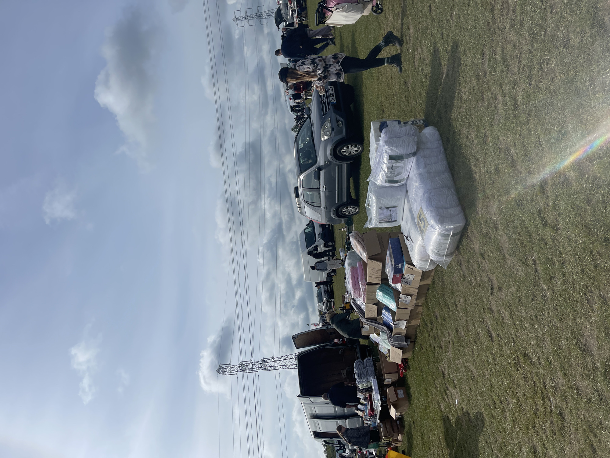 The image shows an outdoor market scene in Sevenoaks, United Kingdom, under a partly cloudy sky. Power lines run across the upper left of the frame. Several cars and vans are parked in a row, acting as stalls for vendors selling various goods. Boxes and packaged items, including bedding, are displayed in front of the vehicles on a grassy field. People are browsing the market, and a few are visible interacting with the merchandise. The scene appears to be a car boot sale or similar open-air market event.