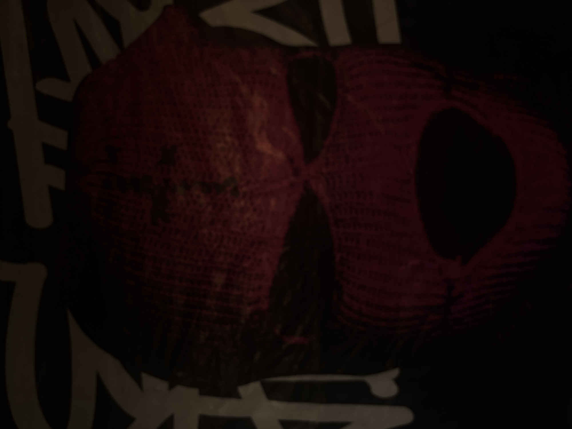The image is a close-up, blurry shot of a red knitted ski mask with black eye holes and a gaping mouth opening. The mask appears to be part of a garment or design, possibly a t-shirt, with abstract white lettering visible in the background. The lighting is dim, casting shadows and obscuring fine details. The overall impression is dark and slightly ominous. The location is Unknown, Unknown.