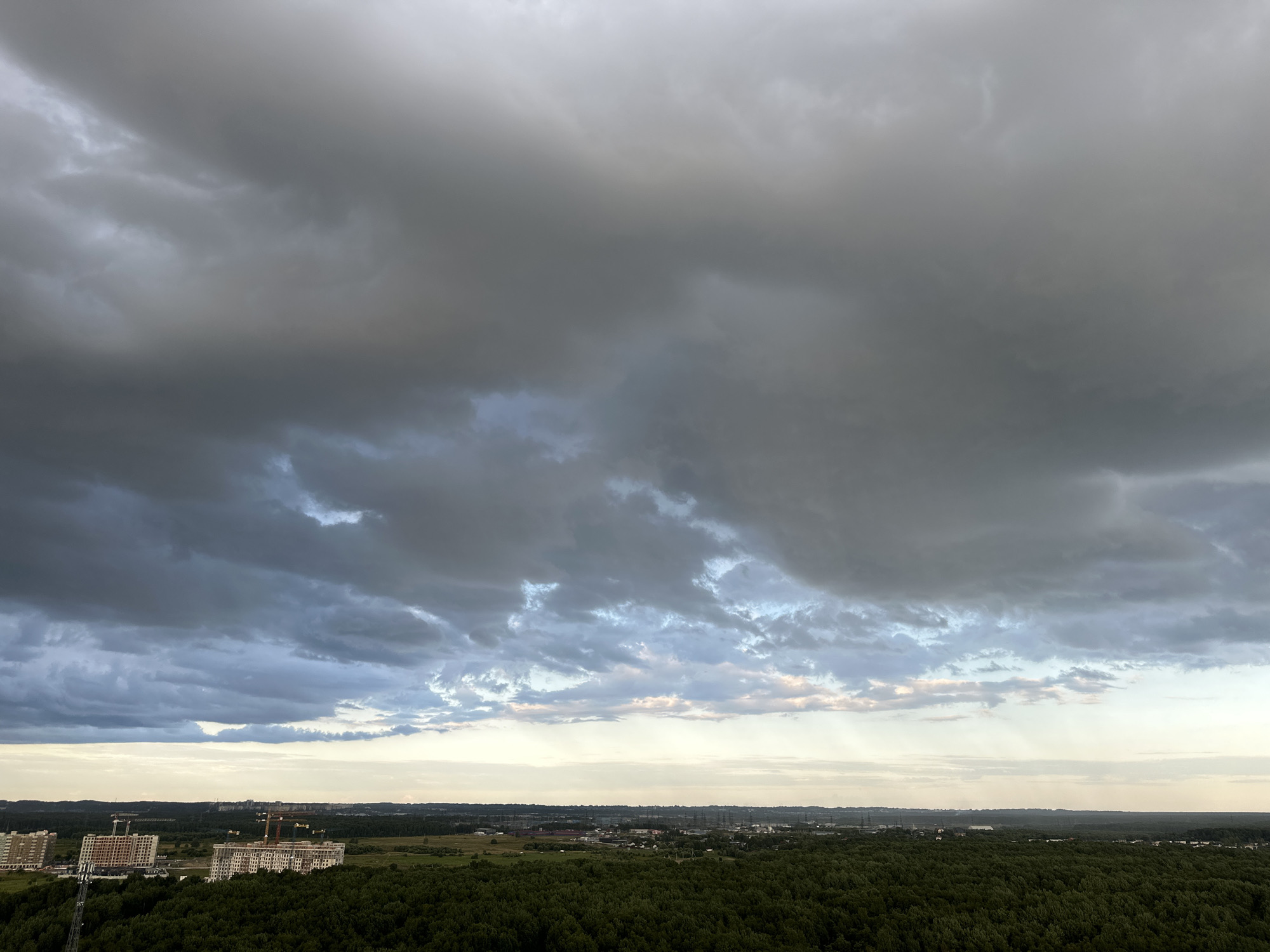 The image depicts a wide-angle aerial view of a suburban landscape under a dramatic, overcast sky, likely at dusk. The sky is filled with heavy, dark grey clouds with lighter patches and hints of blue and pink peeking through, suggesting the presence of sunlight trying to break through or the fading light of day. In the mid-ground, a dense, dark green forest stretches horizontally across the frame. On the edge of the forest and beyond, a developing urban area is visible, characterized by several tall, multi-story buildings, some appearing to be under construction with visible cranes. The buildings are a light color, contrasting with the dark forest and the stormy sky. The overall environment appears to be a mix of natural landscape and ongoing urban development. The location in Dnp Ryzhiki, Russia, is implied, but not visually confirmed within the image itself. There are no people, animals, or vehicles clearly discernible in the image.