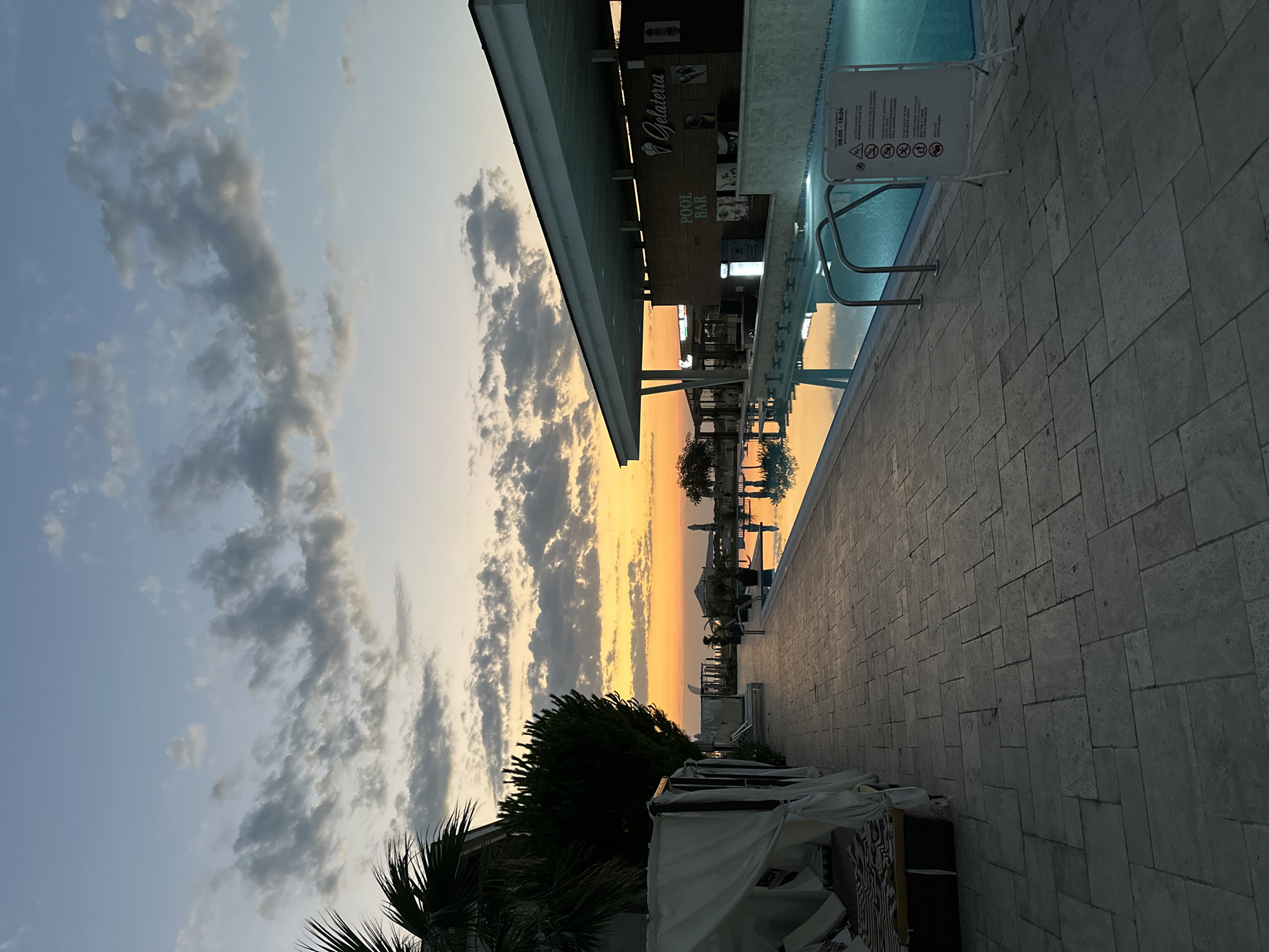 The image depicts a resort poolside at sunset in Sozopol, Bulgaria. The sky is a vibrant gradient of orange, yellow, and blue, with scattered clouds. In the foreground, a stone patio lines a rectangular swimming pool filled with turquoise water. The pool has metal railings leading into it. To the right of the pool is a modern, open-air pool bar and gelateria, with "POOL BAR" and "Gelateria" signage visible. Several chairs and tables are set up under the covered bar area. To the left of the patio, there are white cabanas with seating inside. Palm trees and manicured hedges line the edge of the property. In the distance, the sea is visible, meeting the horizon. The scene is tranquil and suggests a resort atmosphere. There are no people visible in the image.