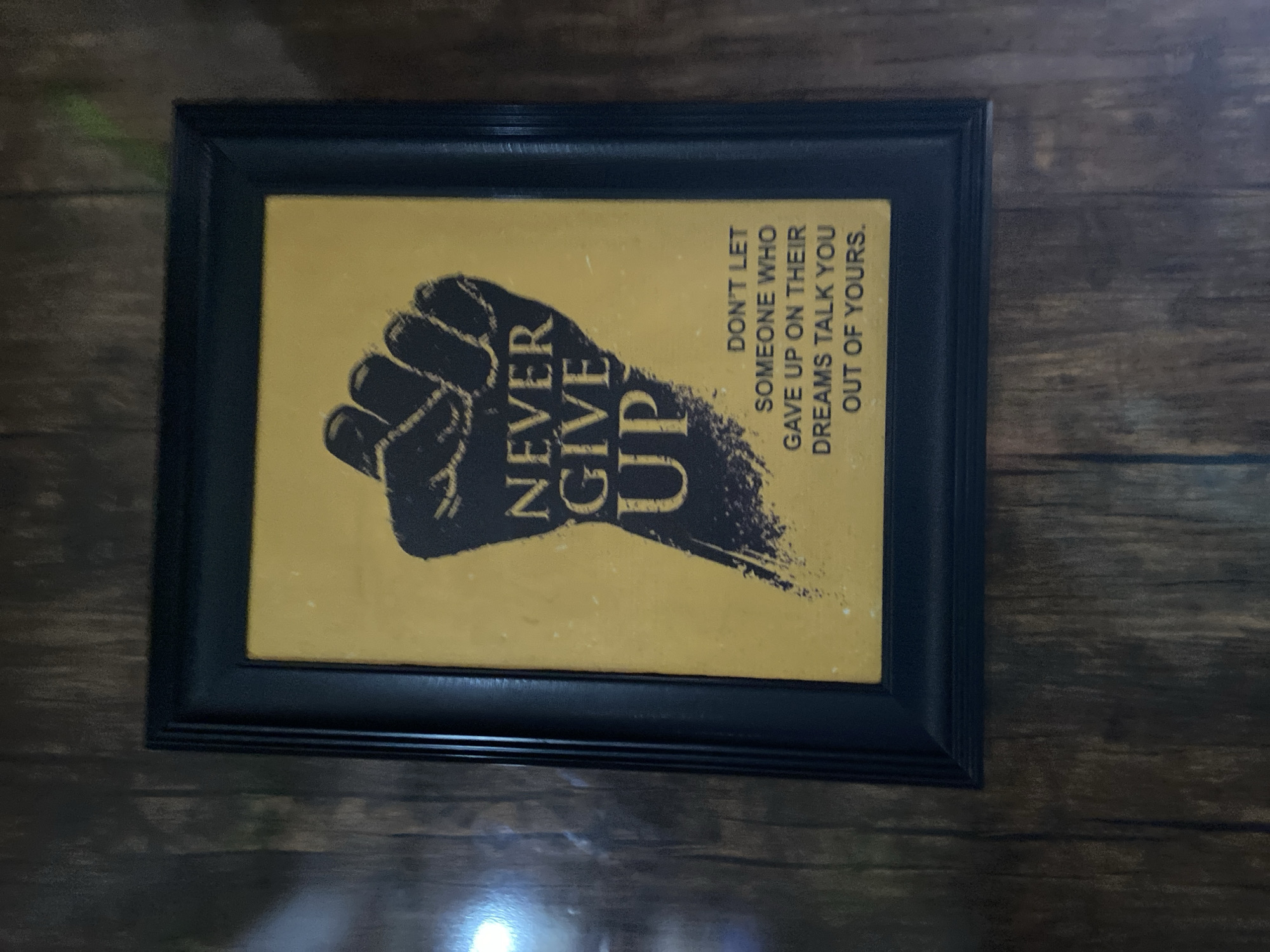 The image shows a framed poster hanging on a wooden wall. The poster has a yellow background with a black fist graphic and the words "NEVER GIVE UP" written vertically. To the right of the fist, the text reads "DON'T LET SOMEONE WHO GAVE UP ON THEIR DREAMS TALK YOU OUT OF YOURS." The frame is black. The wooden wall has a dark brown color with a horizontal grain pattern. The location in the city of Lokoja, Nigeria is not discernible from the image.