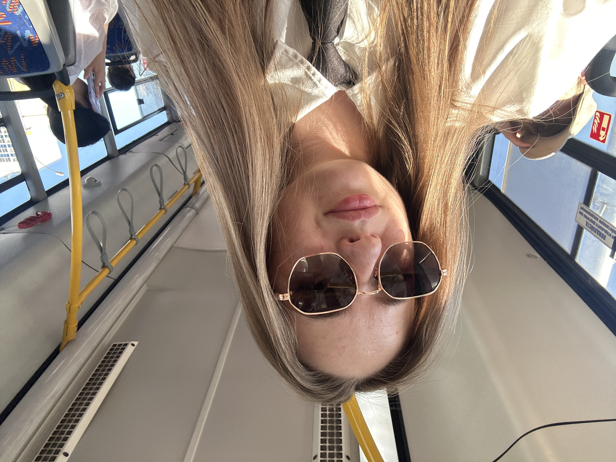 The image, taken from a low angle and rotated, shows a young woman inside a public transportation vehicle, likely a bus or tram, based on the presence of yellow handrails and windows. The woman has long, blonde hair that falls around her face. She is wearing large, gold-rimmed sunglasses. Her expression is neutral, and her head is tilted. She is wearing a white collared shirt with a dark tie visible.
The interior of the vehicle is visible, with gray seats and windows showing a bright, possibly sunny, day outside. A yellow pole is a prominent feature in the foreground. There is a red sandal visible on the floor in the upper left quadrant of the image. A small red sticker with a white symbol is visible on the window. There is some text on a sign in the upper right, but it is largely unreadable. The overall lighting suggests it is daytime. The image appears to be a selfie, taken with the phone held upside down.
