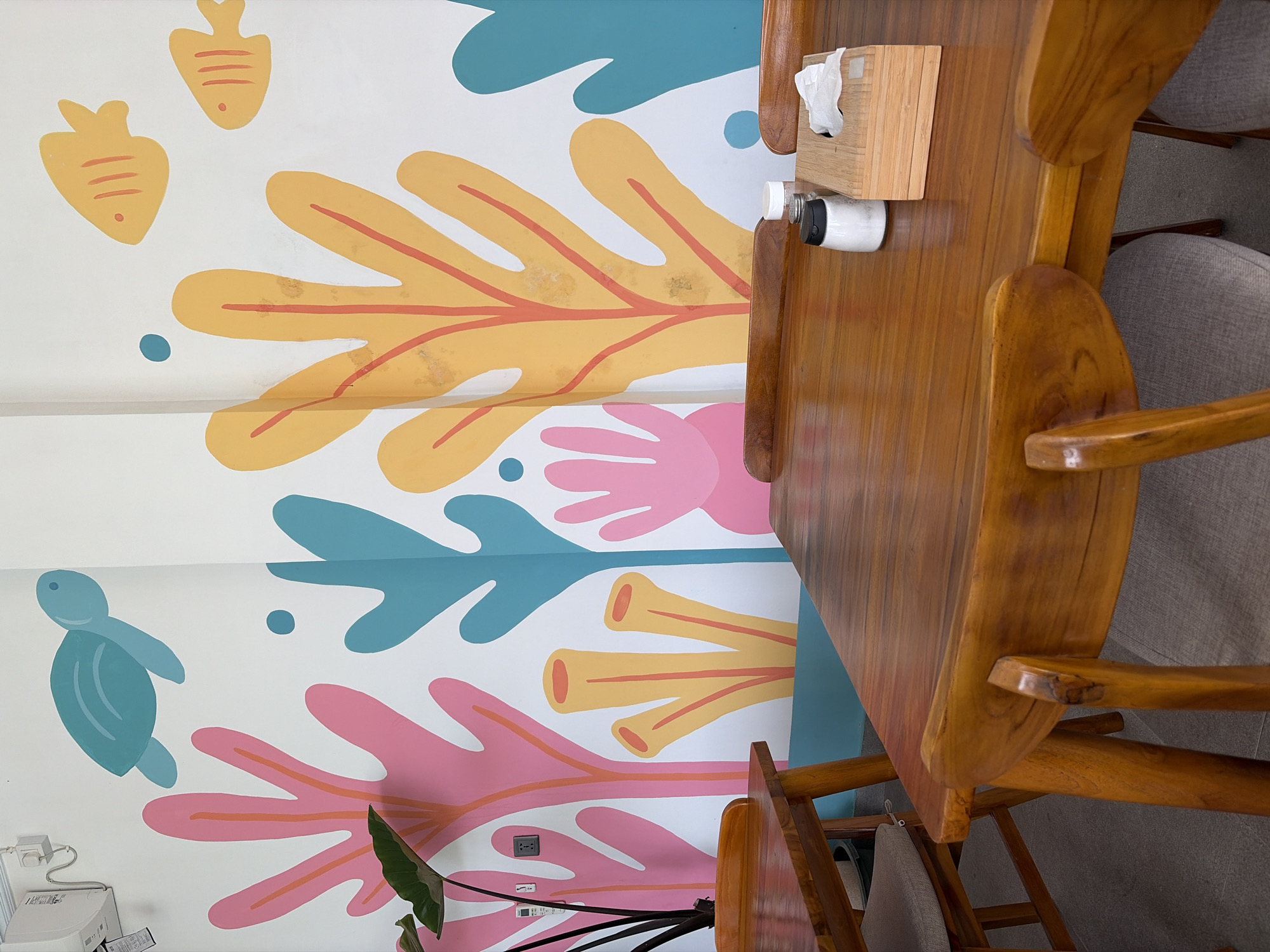 This image depicts a restaurant interior with a vibrant, underwater-themed mural on the wall. The mural features stylized representations of marine life such as a blue turtle, yellow fish, and colorful coral-like shapes in pink, yellow, and teal. In the foreground, two wooden tables are visible, set with chairs. One table has a napkin dispenser, a salt shaker, and a pepper shaker on it. A large green plant is also present in the left side of the frame. The overall impression is a cheerful and casual dining environment. The location is specified as a restaurant in Kubu, Indonesia. There are no people visible in the image, and no actions or interactions are taking place.