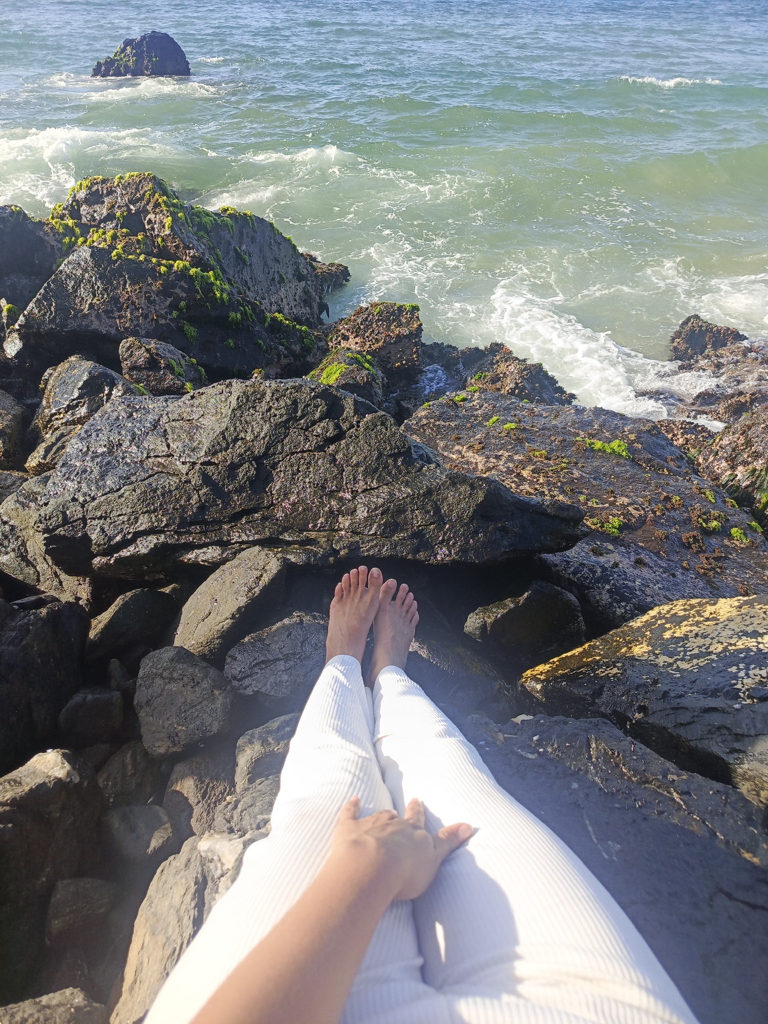 A first-person perspective shot shows a person sitting on a rocky shore with their legs extended towards the ocean. The person is wearing white ribbed pants and their bare feet are visible. Their left hand rests on their left thigh. The rocks are dark and appear wet, with patches of green moss on some of them. The ocean water is a pale green and there are waves crashing against the rocks. In the distance, a lone rock juts out of the water. The scene appears to be a natural, undeveloped coastal area.
