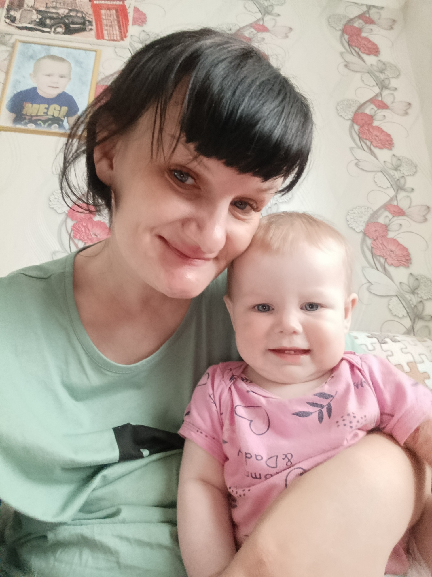 A woman and a baby are indoors, presumably in a home located in Фоки, Russia. The woman has dark hair with bangs and is wearing a light green t-shirt with a dark sleeve underneath. She is smiling and looking at the camera while holding the baby close. The baby has light blonde hair and is wearing a pink onesie with a heart and leaf design on it. The baby is also smiling and looking at the camera. Behind them, on the wall, there is wallpaper with a floral pattern and a framed picture of a young child in a blue shirt. The background also includes a picture of a car and a telephone box. The furniture in the room seems to be a pillow.