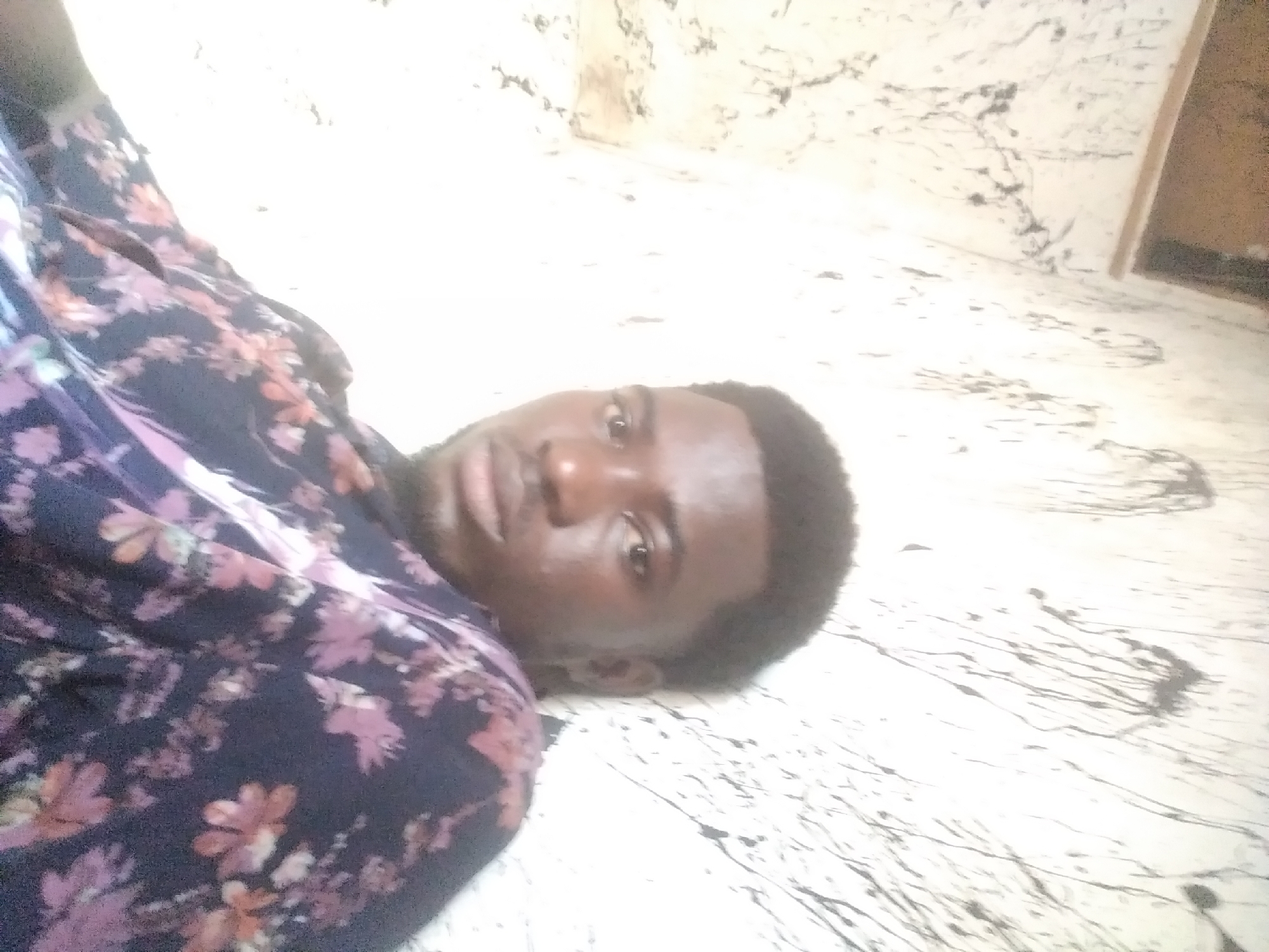 The media file shows a person lying on their back on a patterned floor. The person is wearing a dark-colored shirt with a floral pattern in shades of pink, purple, and white. They have dark hair and are looking towards the camera with their eyes open. The floor has a white base with dark, abstract splatters and lines, suggesting a marble or tiled surface. In the background, there's a hint of a wall and a wooden frame, possibly a doorway or window. The lighting appears to be natural, coming from the side, creating some shadows and highlights on the person's face and the floor. The overall impression is that of a casual, personal photo taken indoors. The location context provided is Jalingo, Nigeria, but there are no specific visual cues in the image to confirm this. There is no visible text in the media file.