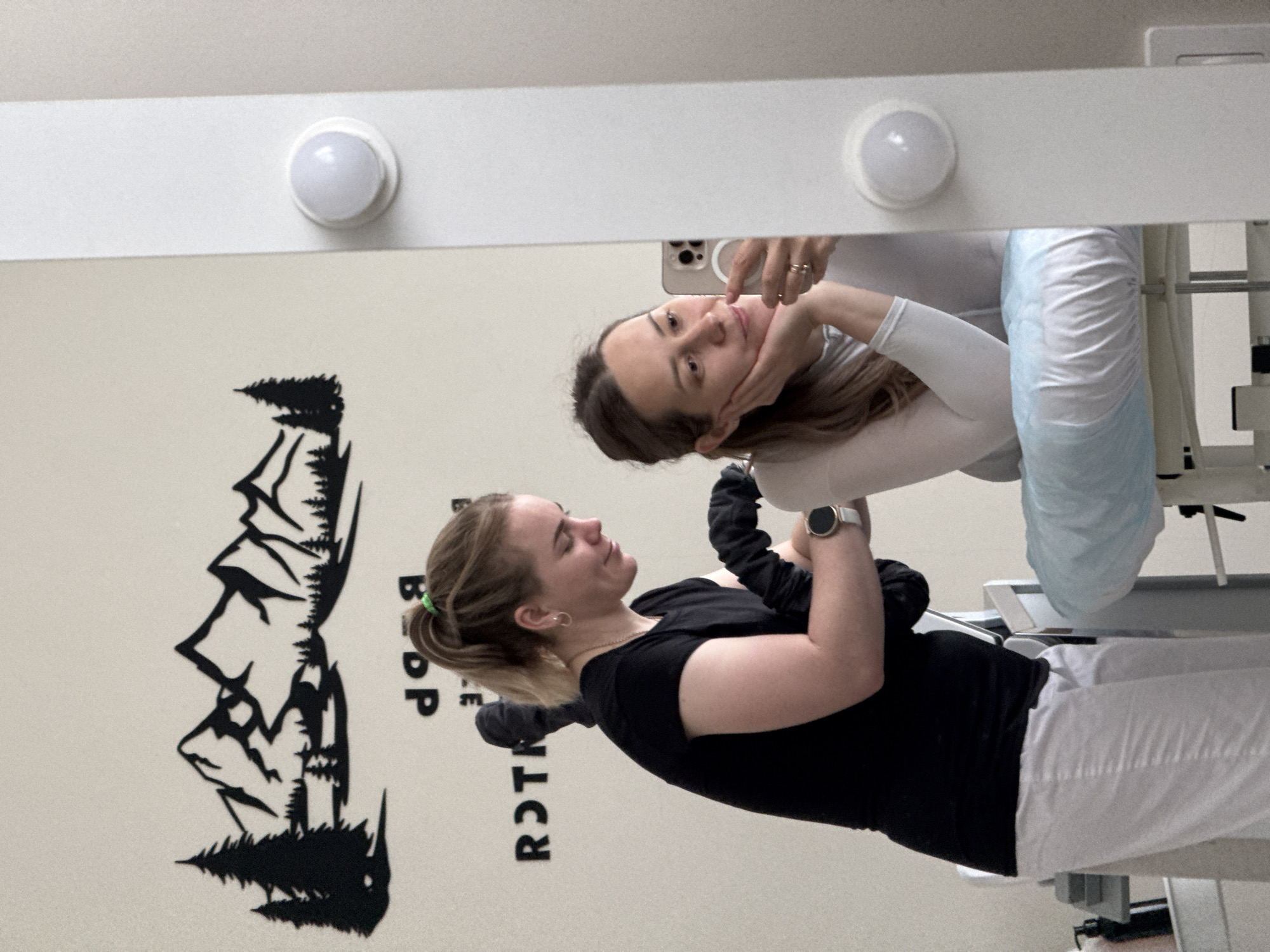 Two women are captured in an indoor setting, likely a beauty or cosmetic studio in Chaikovsky, Russia. The image is taken from the perspective of one woman who is taking a selfie in a large mirror.
In the mirror's reflection, a woman with long, light brown hair, wearing a white long-sleeved top, is seen holding a smartphone (identifiable as an iPhone) to capture the image. She rests her hand gently under her chin and has a subtle, friendly smile. A ring is visible on her left hand.
Directly in front of the mirror, the second woman is seated or reclined on what appears to be a treatment bed, partially covered by a light blue disposable sheet. She has blondish-brown hair tied in a ponytail with a green hair tie. She wears a black short-sleeved top and white pants, and a smartwatch is visible on her left wrist. She looks towards the mirror, smiling calmly.
The background wall behind the second woman features a large black decal depicting silhouetted mountains and pine trees. Visible text on the wall, oriented vertically, includes "B DP" and "ROTЯ". The overall mood of the scene is relaxed and friendly, suggesting a casual moment between the two women.