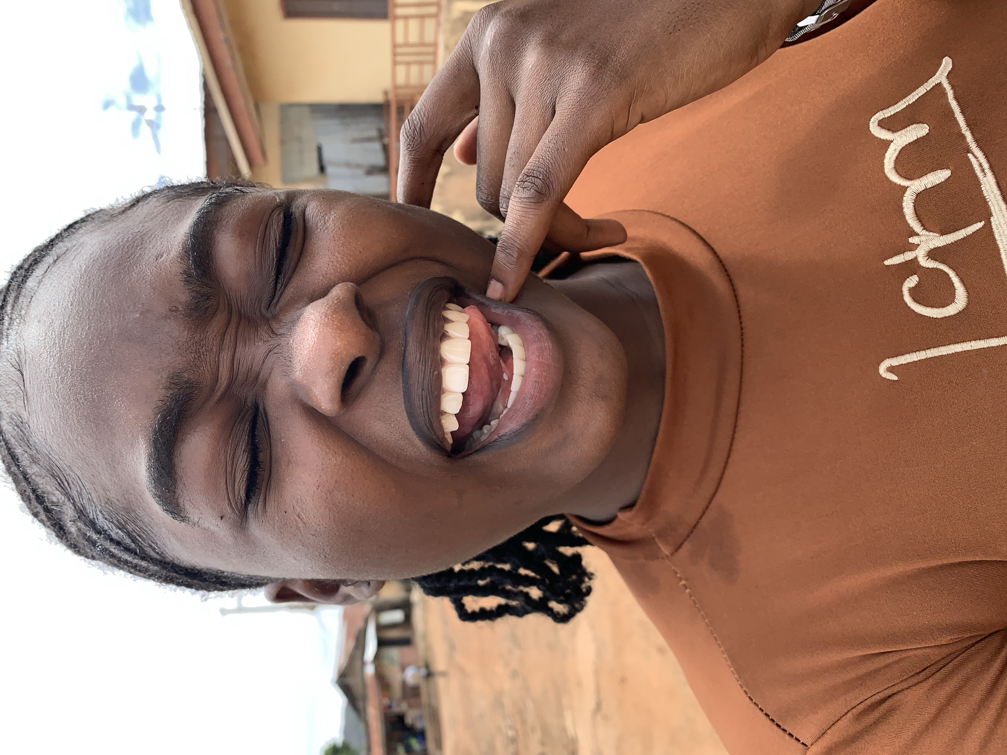 A close-up portrait in Rafin Par, Nigeria, features a young Black woman with a dark complexion, positioned center-frame against an outdoor background. Her eyes are tightly closed and squinted, creating prominent wrinkles on her forehead, around her eyes, and on her nose. Her mouth is wide open, revealing white teeth and a pink tongue slightly extended and curled towards her left. Her right hand (viewer's left) has the index and middle fingers pressing against her lower lip and chin area, further exaggerating the open mouth. Her hair is styled in neat flat braids visible along her hairline, with some longer braids falling on her right side (viewer's left). She is wearing a rust-brown or burnt orange long-sleeved, high-neck top. The embroidered text "I am" in a light, possibly cream-colored script, is partially visible on the lower right of her shirt. The background consists of reddish-brown dirt ground in the foreground. Distant, out-of-focus buildings are visible: on the left, a utility pole and simple structures with a blurred figure; on the right, a light yellow single-story building with a reddish-brown roof, a dark window shutter, and a railing. The sky above is bright and overcast with subtle cloud formations.