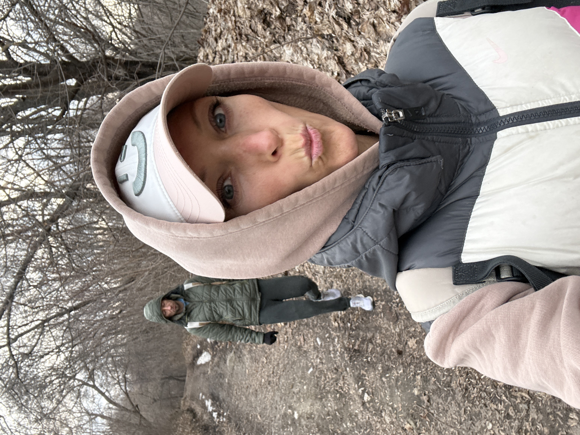 The image shows a selfie of a woman with blue eyes and a slight pout. She is wearing a white and grey Nike jacket with a pink hooded sweatshirt underneath and a white visor with a green design. Behind her, another person is walking on a path in a wooded area. The location appears to be a park or wooded area in Ulyanovsk, Russia, given the bare trees and autumn foliage.