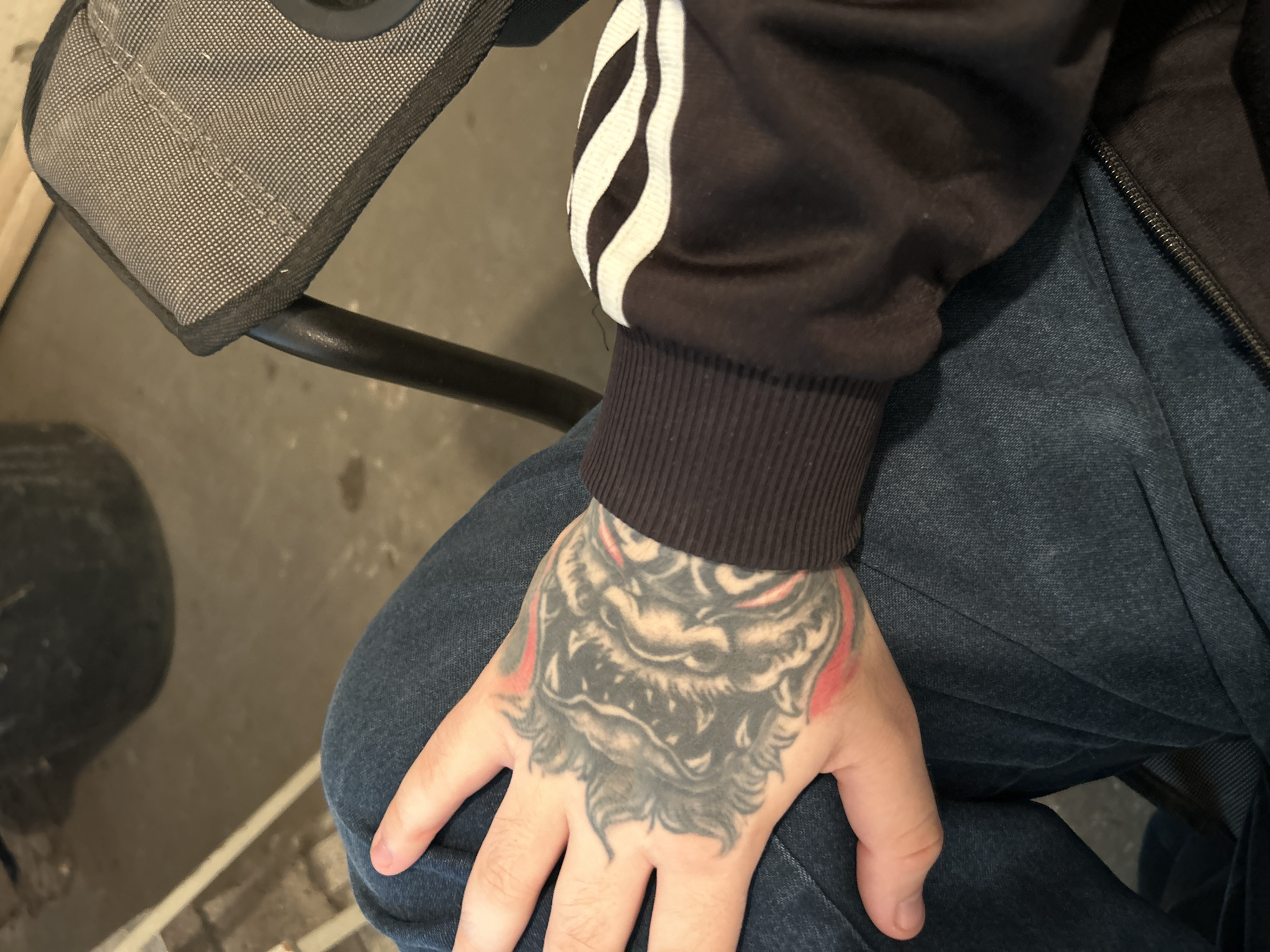 The image shows a close-up of a person's hand with a tattoo of what appears to be a wolf's head. The hand is resting on a pair of dark blue jeans. The person is wearing a dark long-sleeved jacket with white stripes on the sleeves. In the background, part of a gray folding chair and a concrete floor are visible. There is no discernible information in the image that allows for the specific location within Gelsenkirchen, Germany to be identified.