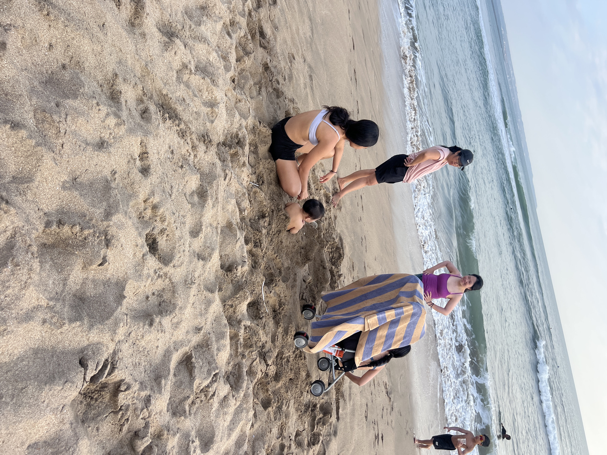 In Kuta, Indonesia, a family enjoys a beach day. The scene unfolds on a sandy shore, with the ocean in the background featuring gentle waves and several surfers. In the foreground, a baby is partially buried in the sand, being observed by a young woman sitting nearby. Another woman stands closer to the ocean, looking out at the water, while a third woman stands with hands on her hips. A stroller with a striped blanket draped over it is positioned amongst the group. A young boy walks near the water's edge, holding a drink. The overall atmosphere is one of relaxed family recreation.