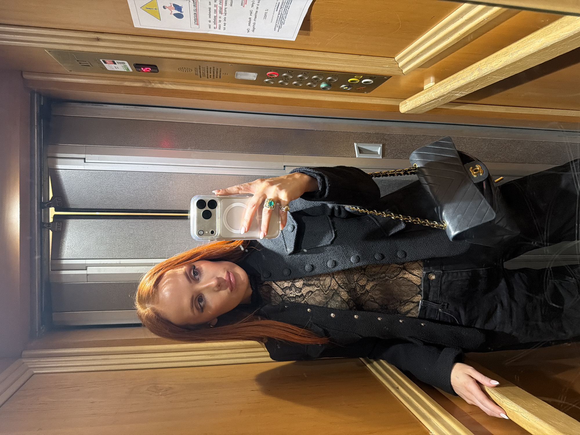 A woman with long red hair is taking a selfie in an elevator. She is wearing a black jacket with buttons, a lace top, and black jeans. She is holding a cell phone in her left hand, and a black quilted purse is visible on her shoulder. The elevator has wooden paneling, a metal door, and a display showing the floor number. There is also a sign with instructions or warnings in the upper left corner of the elevator. The location is likely in Paris, France.