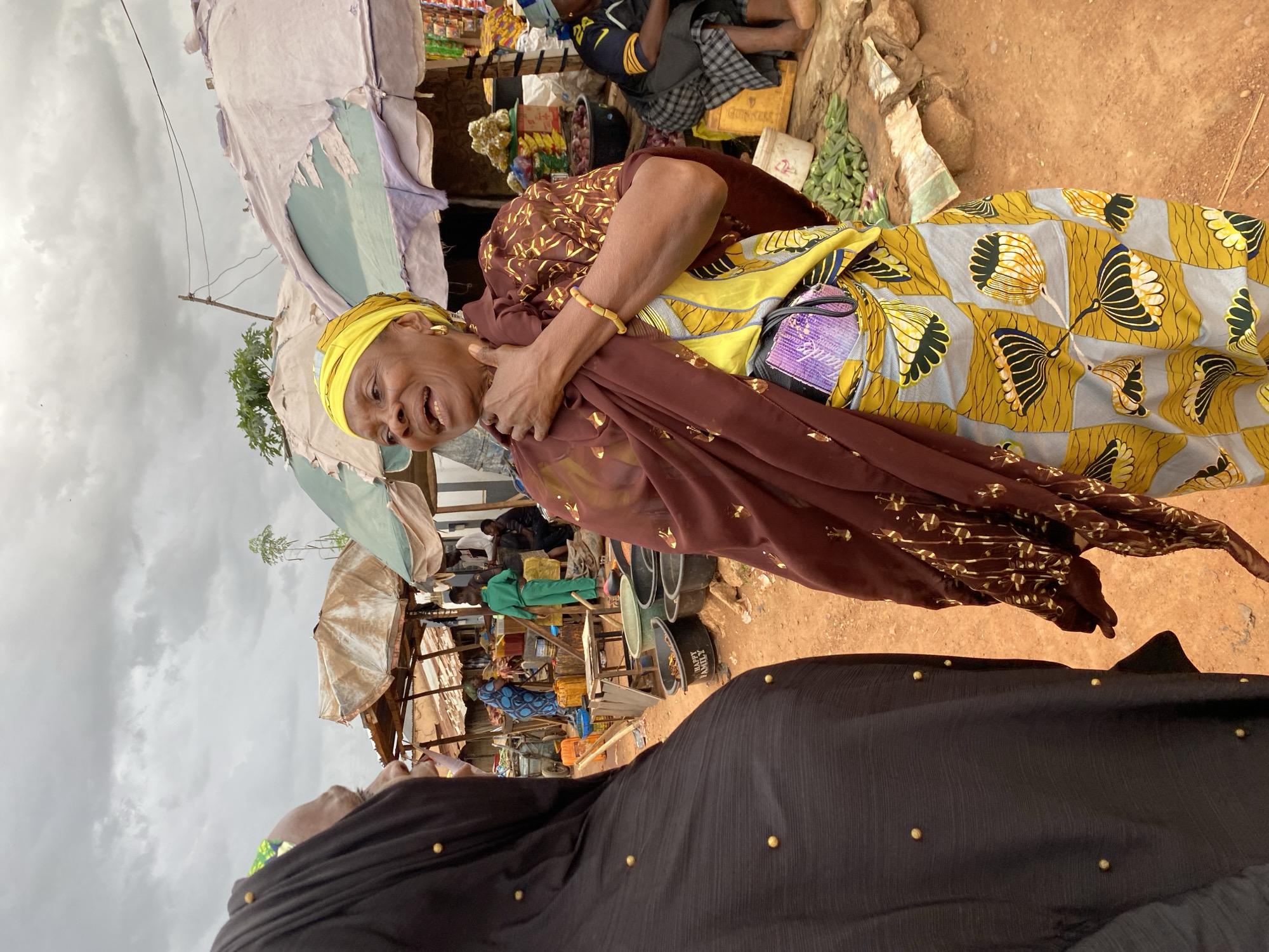 The image depicts an outdoor market scene in Bukuru, Nigeria, under a cloudy sky. In the foreground, two women are engaged in conversation. The woman on the right, facing the viewer, wears a vibrant yellow headwrap, a patterned brown and gold shawl draped over her shoulders, and a colorful yellow, blue, and grey patterned skirt. She has a hand resting on her chest and appears to be speaking animatedly. To her left, another woman is mostly obscured, wearing a black hijab with gold bead detailing.
Behind them, the market is bustling with activity. Stalls are set up with various goods, covered by weathered umbrellas. Several people are visible in the background, including a person in a green tracksuit and others tending to goods and interacting. Various containers, including black basins with the inscription "HAPPY FAMILY", are placed on the ground. Produce, such as okra, is visible near the stalls. The ground is covered in reddish-brown earth, typical of the region. The overall impression is one of a lively, everyday market in a Nigerian city.