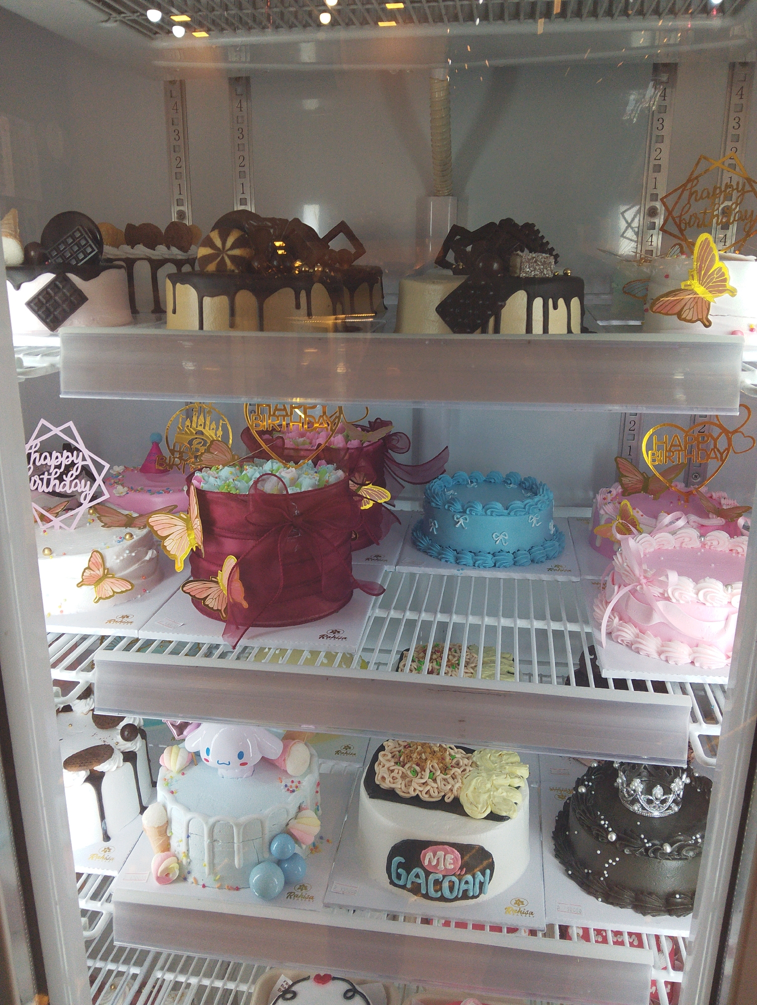 The image shows a refrigerated display case filled with various cakes. The display case is likely located in a bakery or cake shop in Manaruwi, Indonesia.
The cakes are arranged on multiple shelves. The top shelf features several chocolate cakes with decorative toppings, including drizzles and what appear to be chocolate pieces. Some of these cakes also have "Happy Birthday" decorations and butterfly embellishments.
The middle shelf has a variety of cakes. One is a pale pink cake with scattered sprinkles and butterfly decorations. Next to it is a dark red cake adorned with a large bow and butterfly decorations. Further along this shelf, there is a bright blue cake with a piped border and small bow decorations. To the right, a pink cake is decorated with piped frosting and ribbons.
The shelf below displays more cakes. A light blue cake is decorated with a character that resembles Cinnamoroll from Sanrio, along with small ice cream cones and marshmallows. To its left is a white cake with chocolate drizzles. On the right side of this shelf, there is a dark cake topped with a silver crown and pearl-like decorations. Another cake on this shelf has a black rectangle with text that reads "ME GACOAN" in colorful lettering, and is topped with what looks like noodles and light green frosting swirls.
The background of the display case is a light gray, with vertical metal bars and fluorescent lighting visible at the top. The cakes are placed on white cardboard bases. The overall impression is one of a well-stocked and varied selection of cakes.