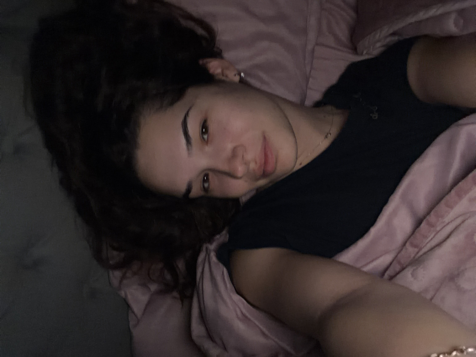 A young woman with dark, curly hair is lying in bed. She is wearing a black t-shirt and has a gold bracelet on her left wrist. She is lying on a light pink bedsheet, with the same color bedsheet on top of her, and the dark wall can be seen on the left. There is no indication or evidence that the location is in Dobrich, Bulgaria.