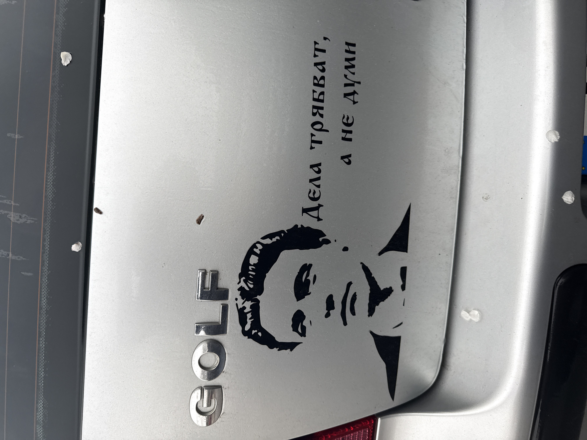 The image shows the back of a silver Volkswagen Golf car. The car has the word "GOLF" badged on the left side. A sticker is placed on the car featuring a black silhouette of a man, positioned below Cyrillic text that translates to "Deeds are needed, not words." The sticker and the image is placed on the rear hatch near the rear tail light on the right side of the car. The scene is likely set in Varna, Bulgaria, due to the Cyrillic text on the sticker. There are various white marks (likely bird droppings) on the silver surface of the car.