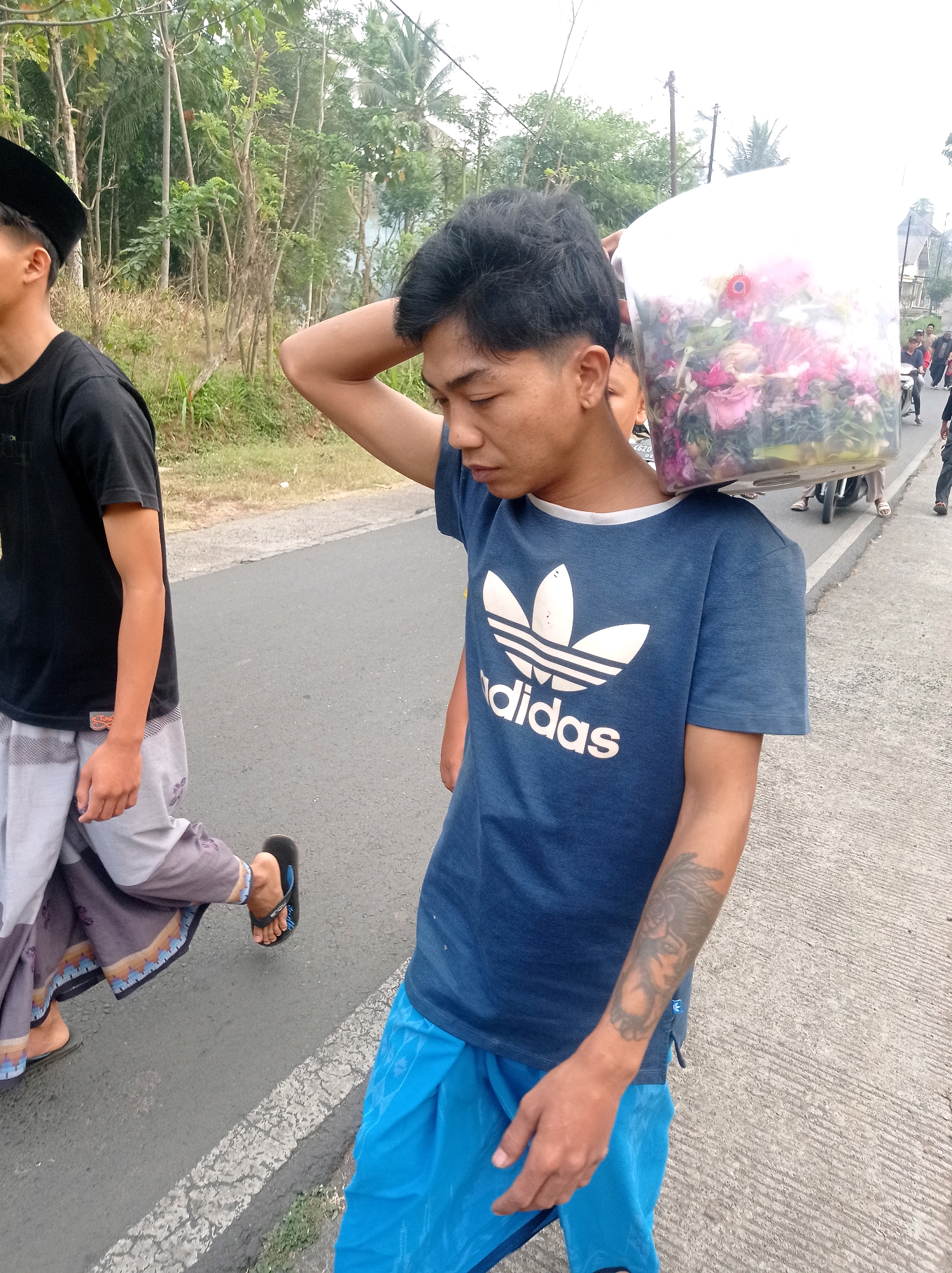 In Pandanmulyo, Indonesia, a young man in a blue Adidas t-shirt walks on a paved road, carrying a clear plastic container filled with pink and red flowers on his left shoulder. He has a tattoo on his right forearm. To his left, another young man in a black t-shirt and a black peci hat walks alongside him. In the background, trees and vegetation line the side of the road. Other people and a motorcycle are visible further down the road. The weather appears overcast.