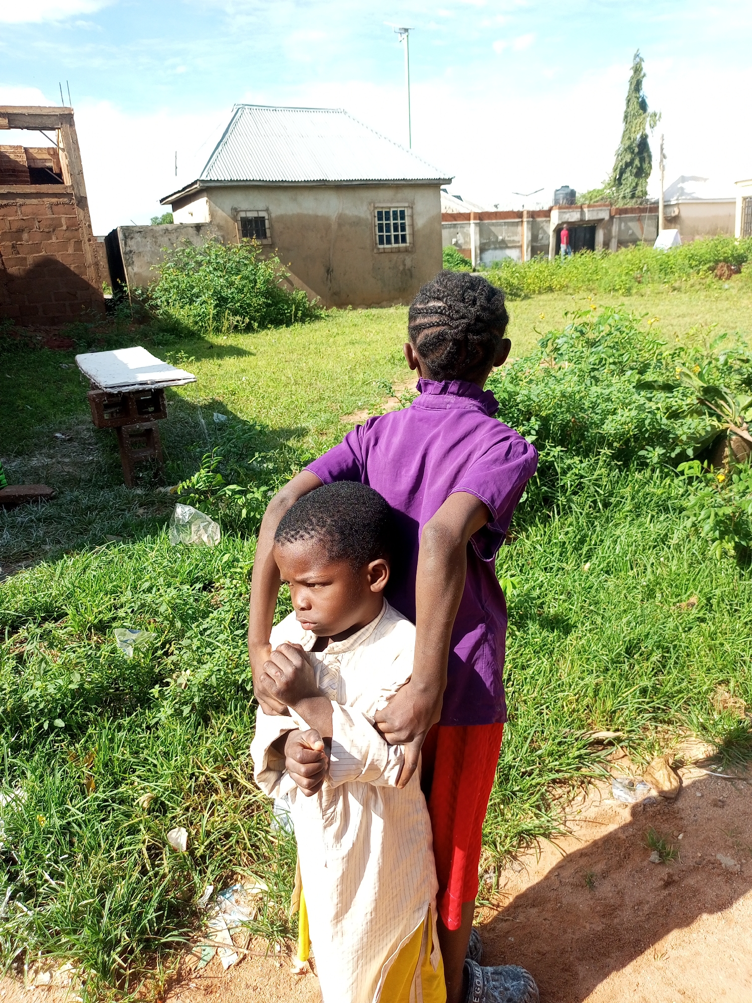 Two young children stand in an outdoor, rural setting. The child in the foreground, facing left, is wearing a light-colored, striped garment and has their arms held by the child behind them. This child has dark hair and appears to be looking down. The child in the background, facing away from the camera, is wearing a purple top and red shorts. They are holding the child in front of them from behind, with their arms around the other child's torso.
The environment consists of green grass, some scattered debris, and a dirt path. In the background, there are several buildings, including a light-colored house with a metal roof and a partially constructed brick building. A tall, thin tree stands in the distance, along with other structures that suggest a residential area. The sky is blue with some white clouds, indicating a daytime scene. The overall impression is a snapshot of everyday life in a non-urbanized area.